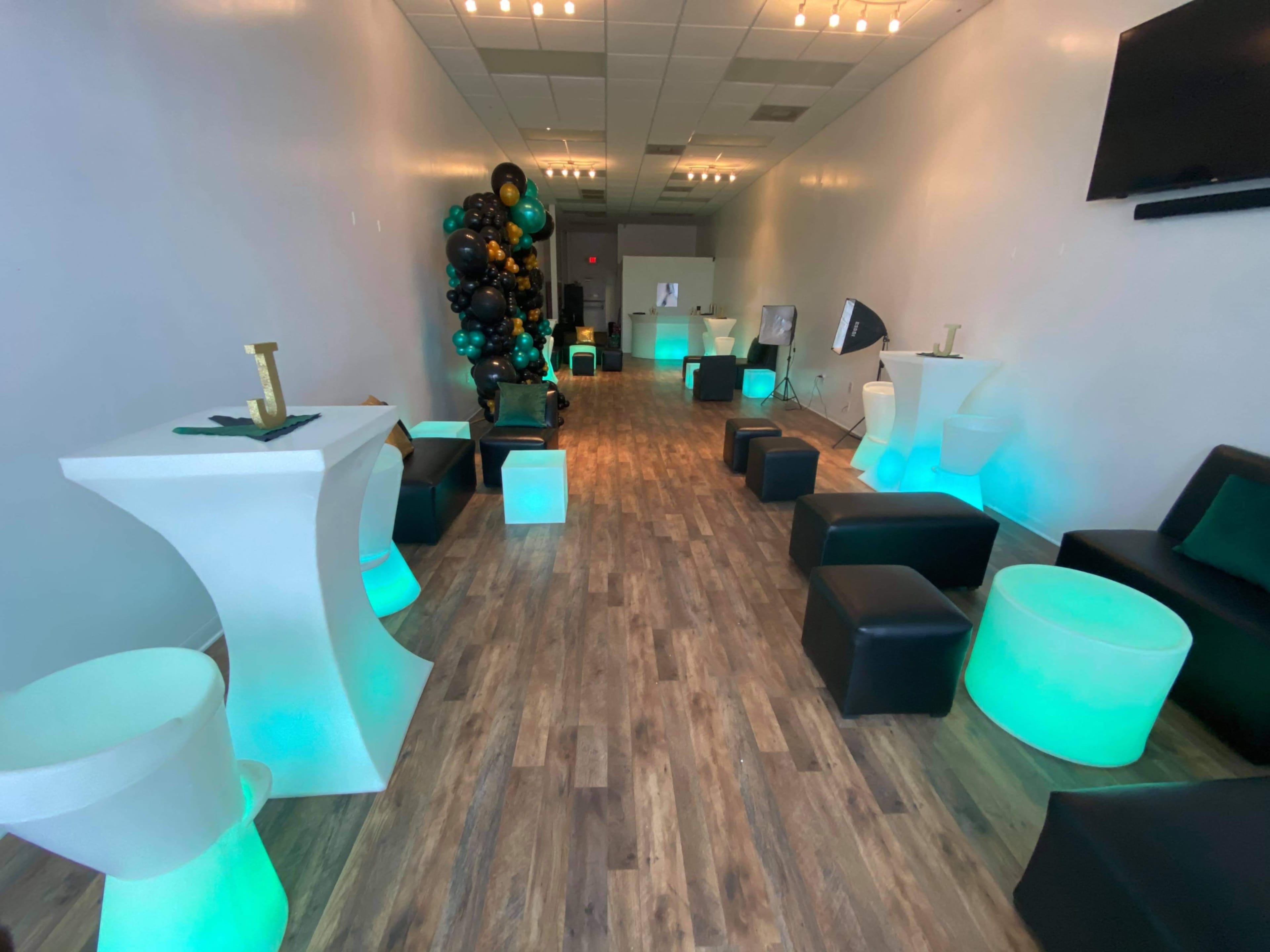 A spacious indoor venue featuring a long, wooden floor, black seating, illuminated green and white tables, and a decorative balloon display in one corner.