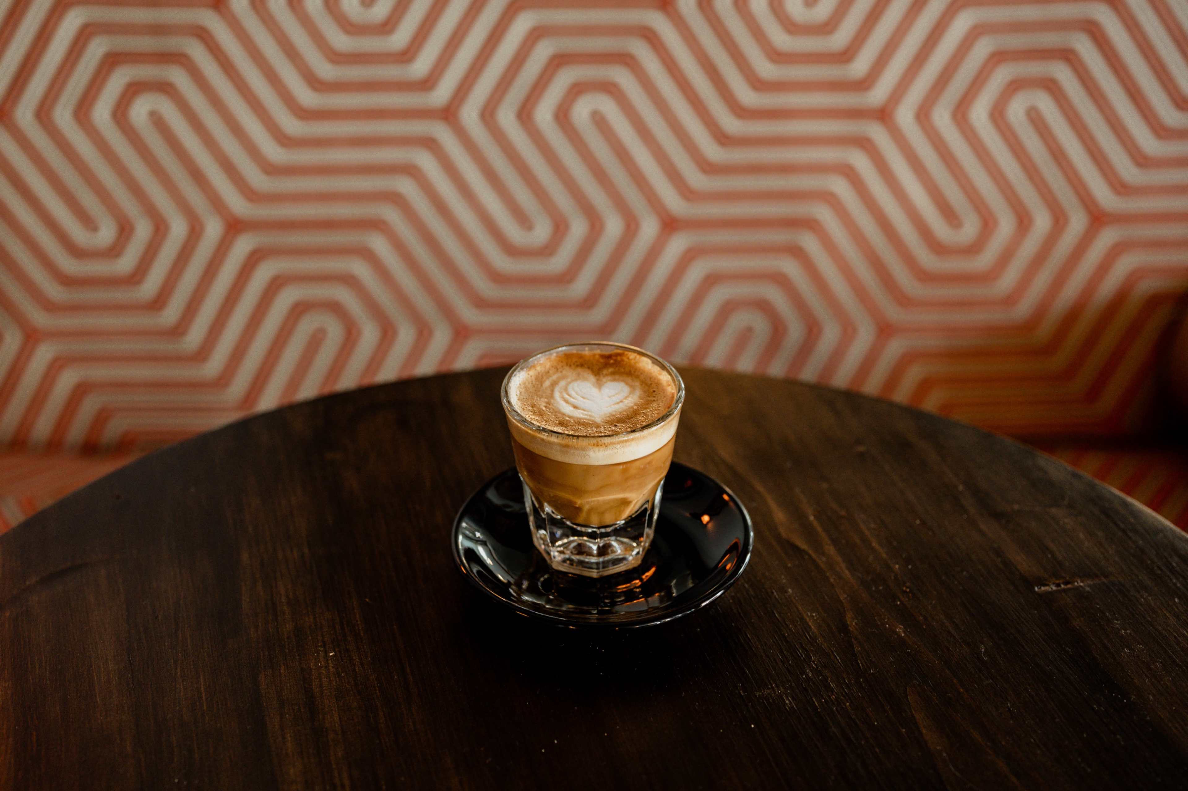 A glass of coffee with a heart-shaped foam design sits on a black saucer atop a wooden table, with a patterned background.
