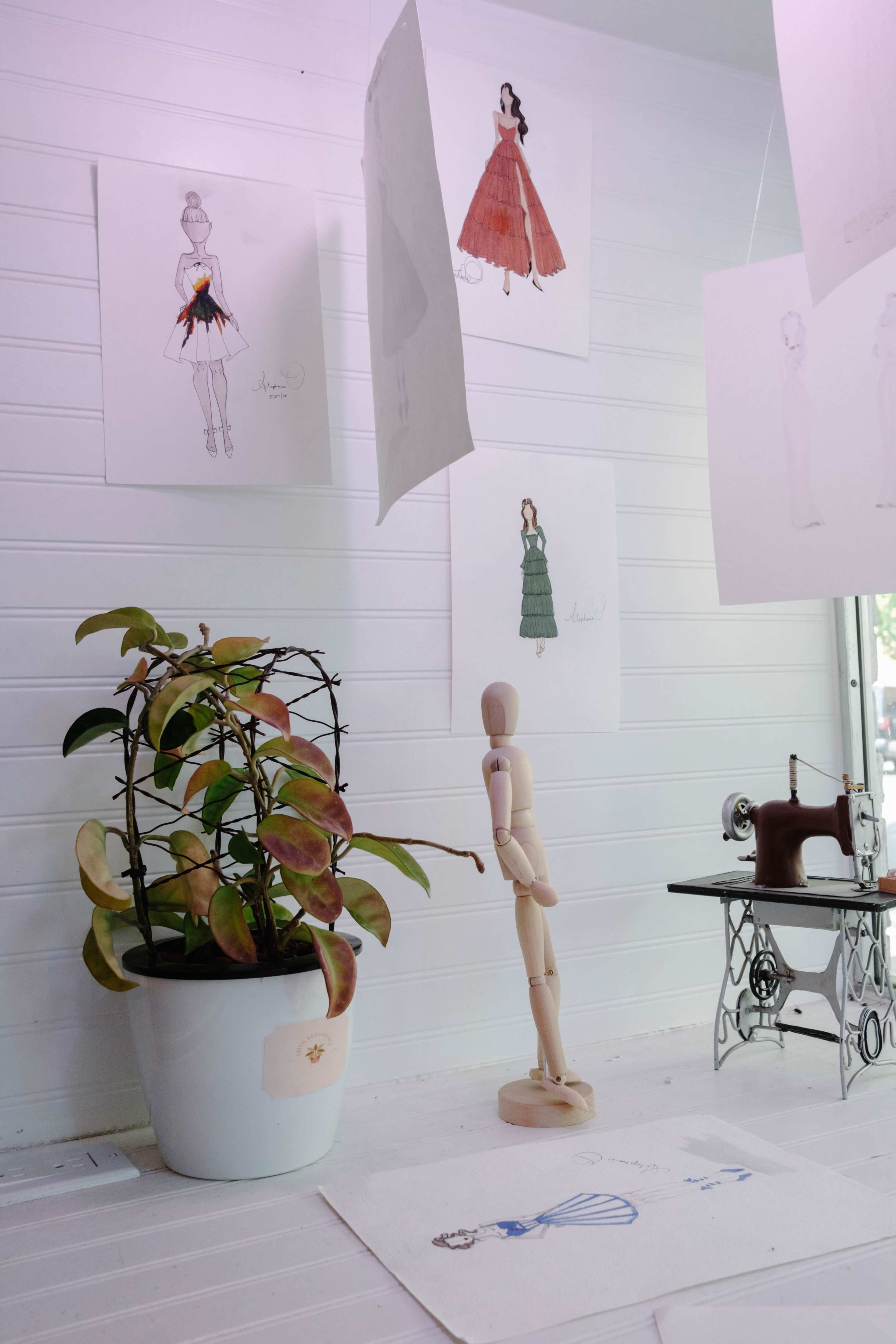 The image shows a bright space featuring fashion sketches hanging on the walls, a potted plant, a wooden mannequin, and a vintage sewing machine.