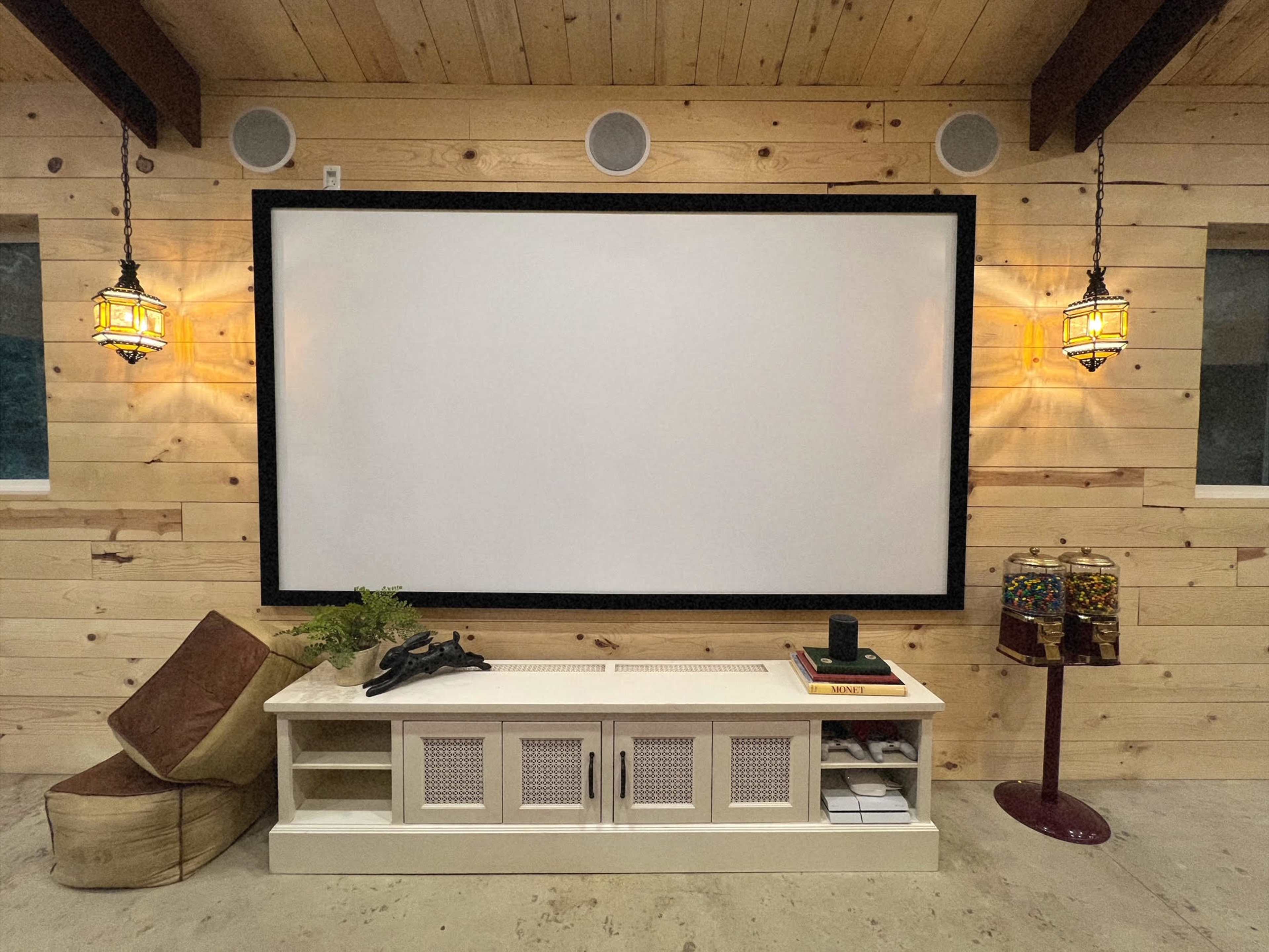 A large, framed projector screen mounted on a wooden wall, flanked by two wall lamps, with a console table and various decorative items beneath it.