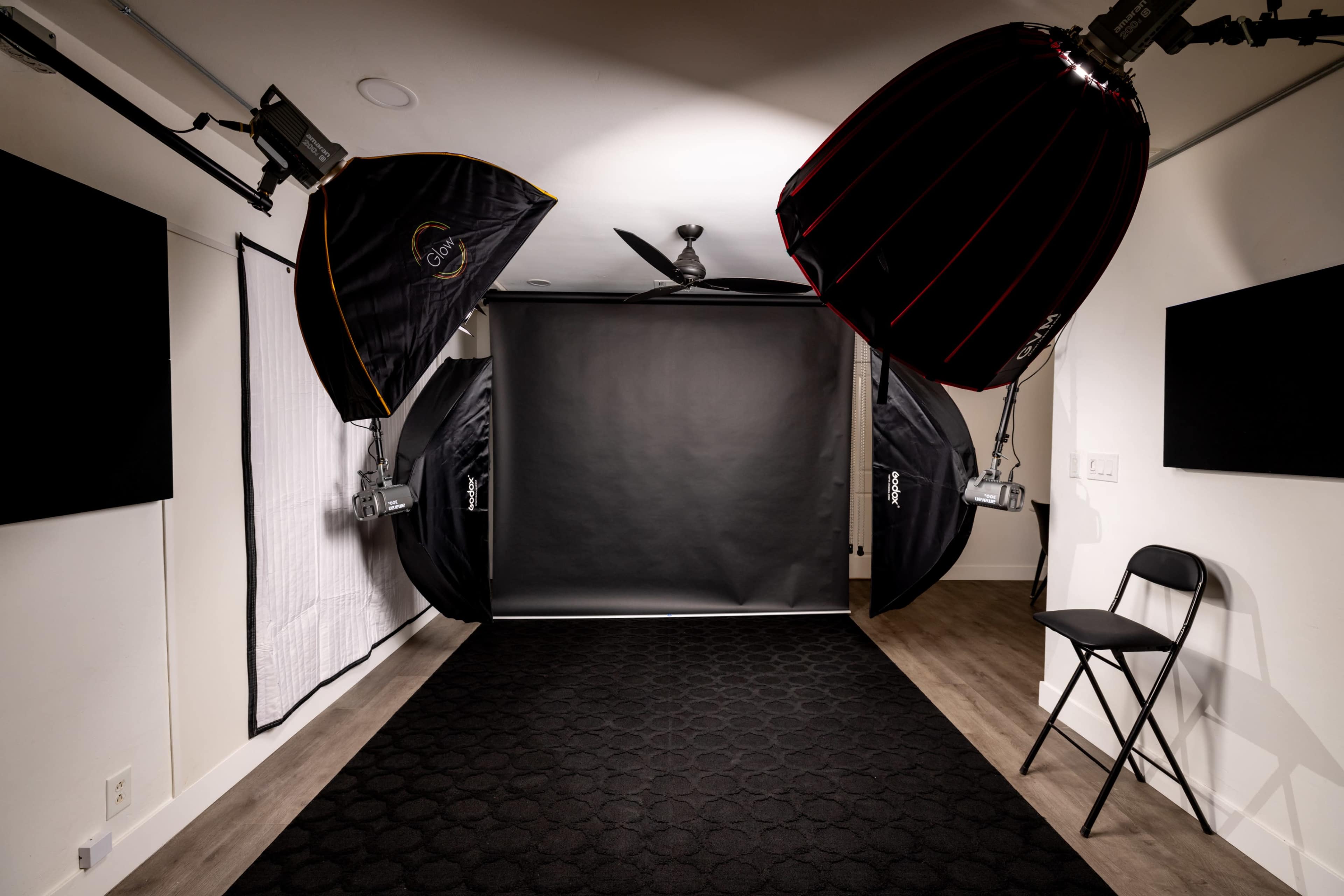 The image shows a photography studio with a black backdrop, two softbox lights, and a folding chair.