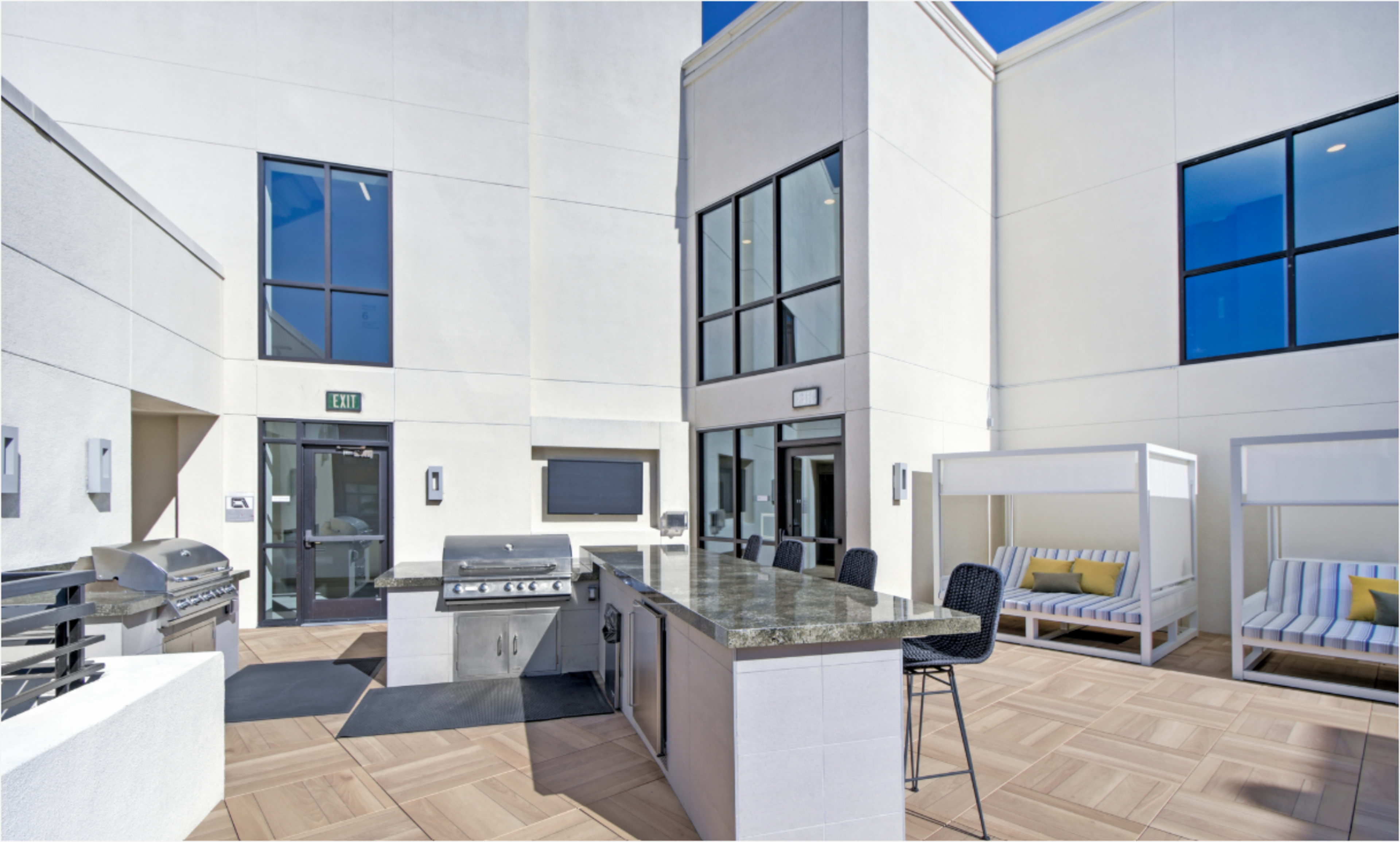 The image shows an outdoor kitchen area featuring a grill, countertop, and seating in a modern space surrounded by white walls and large windows.