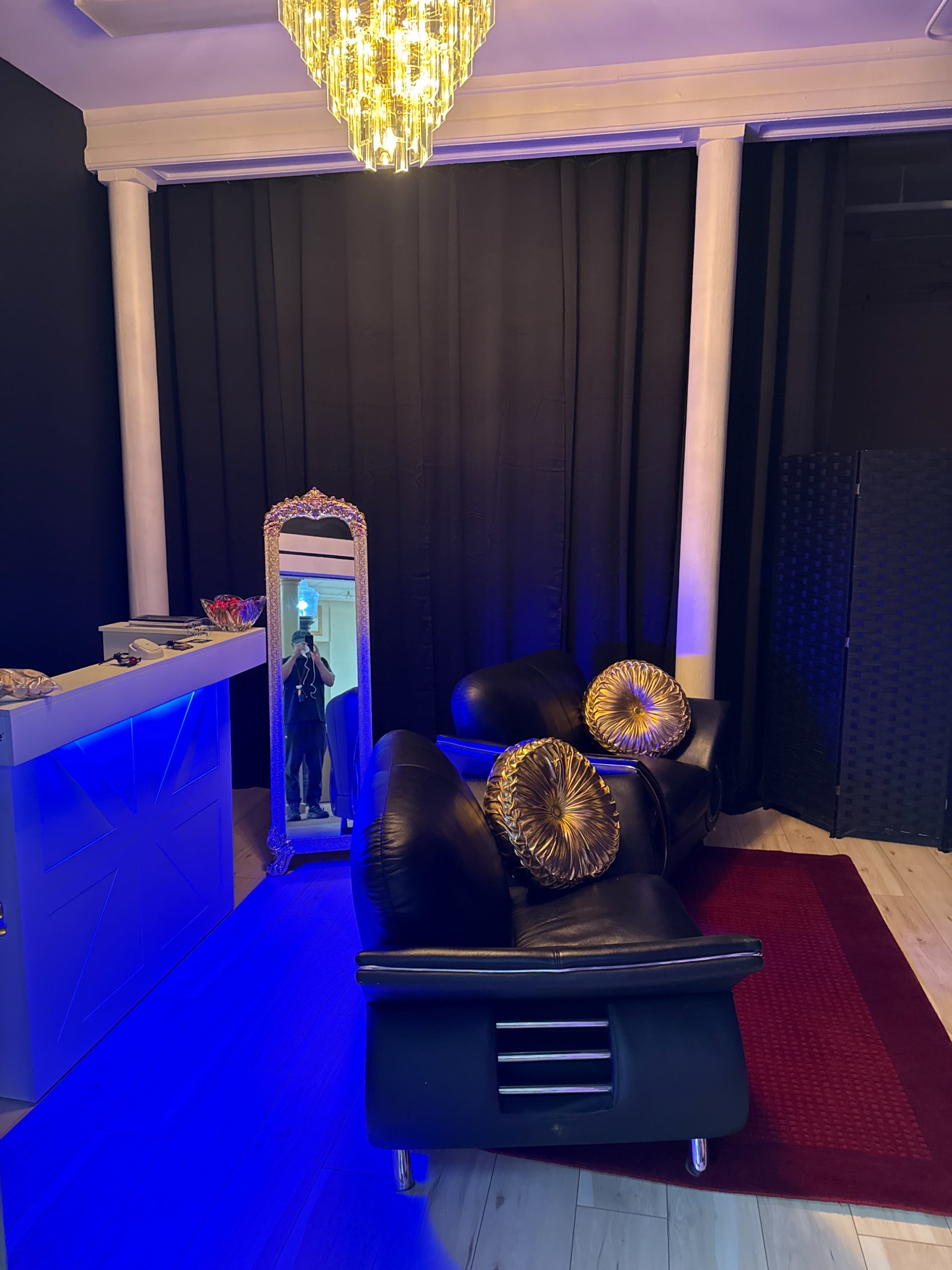 The image features a modern lounge area with a black leather couch, decorative pillows, a tall mirror, and a chandelier, illuminated by blue lighting.