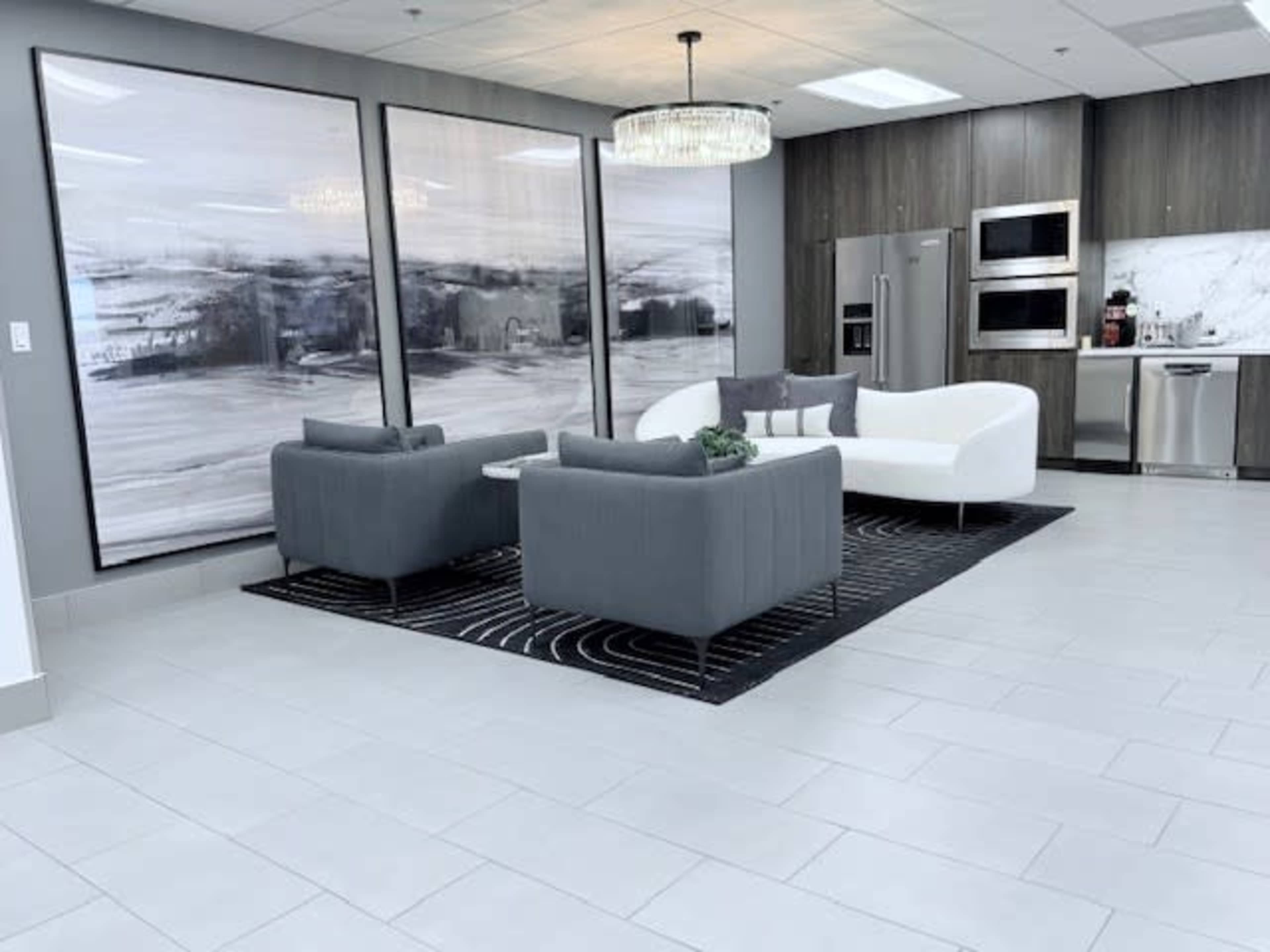 The image features a modern lounge area with two gray armchairs and a white sofa, set on a patterned black and white rug, surrounded by large abstract artworks and stainless steel kitchen appliances.