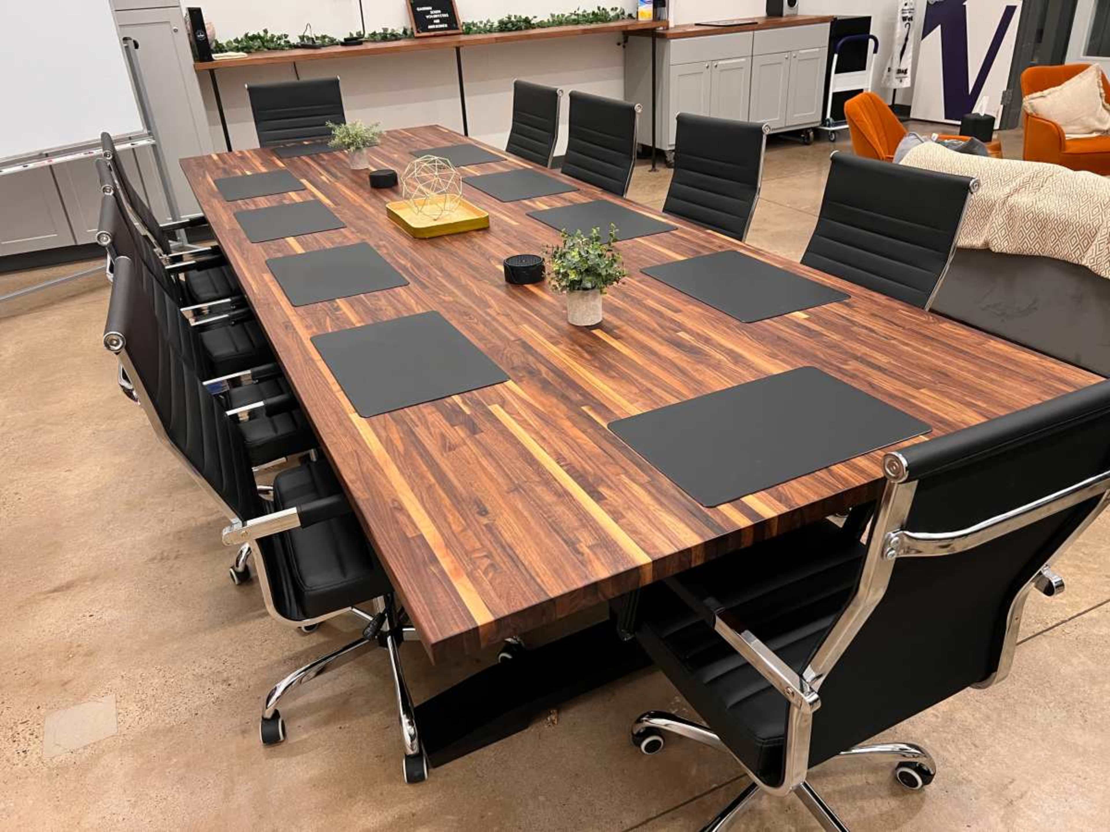 A long wooden conference table with black placemats surrounded by black leather chairs occupies a well-lit meeting room.