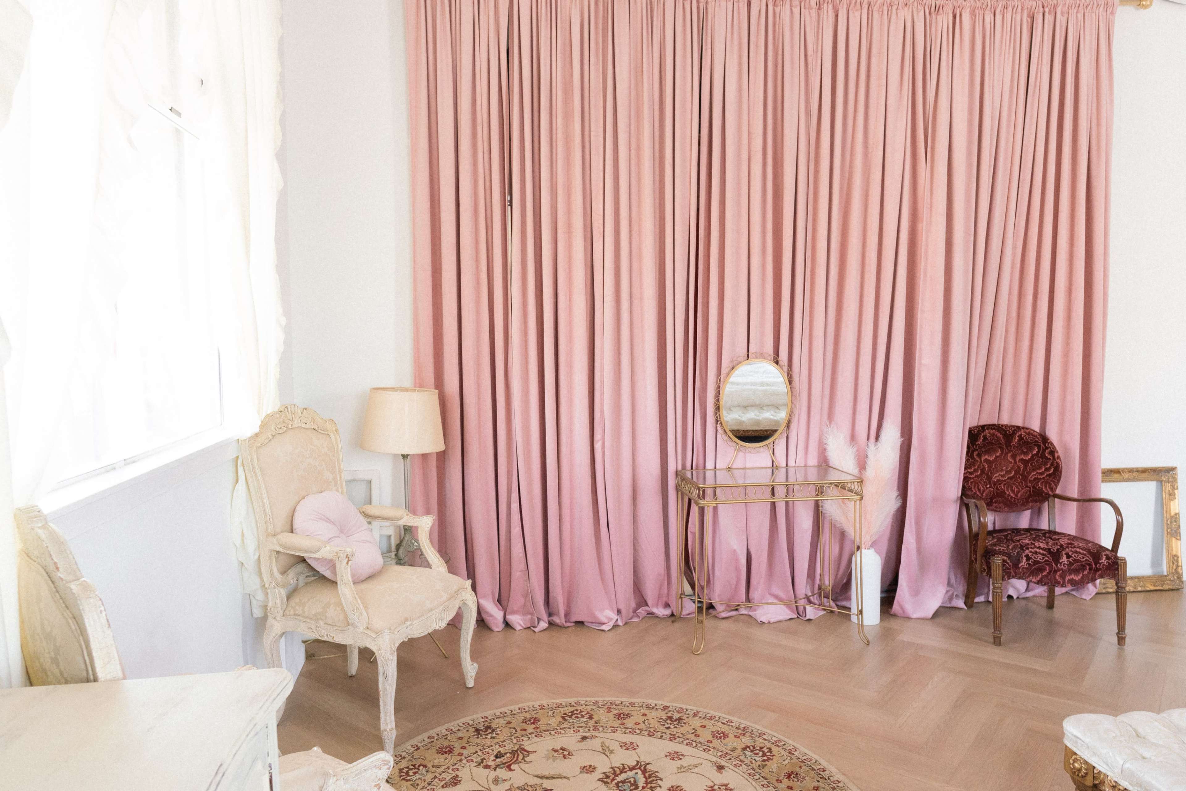 The image shows a room with pink curtains, a mirror on a glass table, two chairs, and a decorative rug on a wooden floor.
