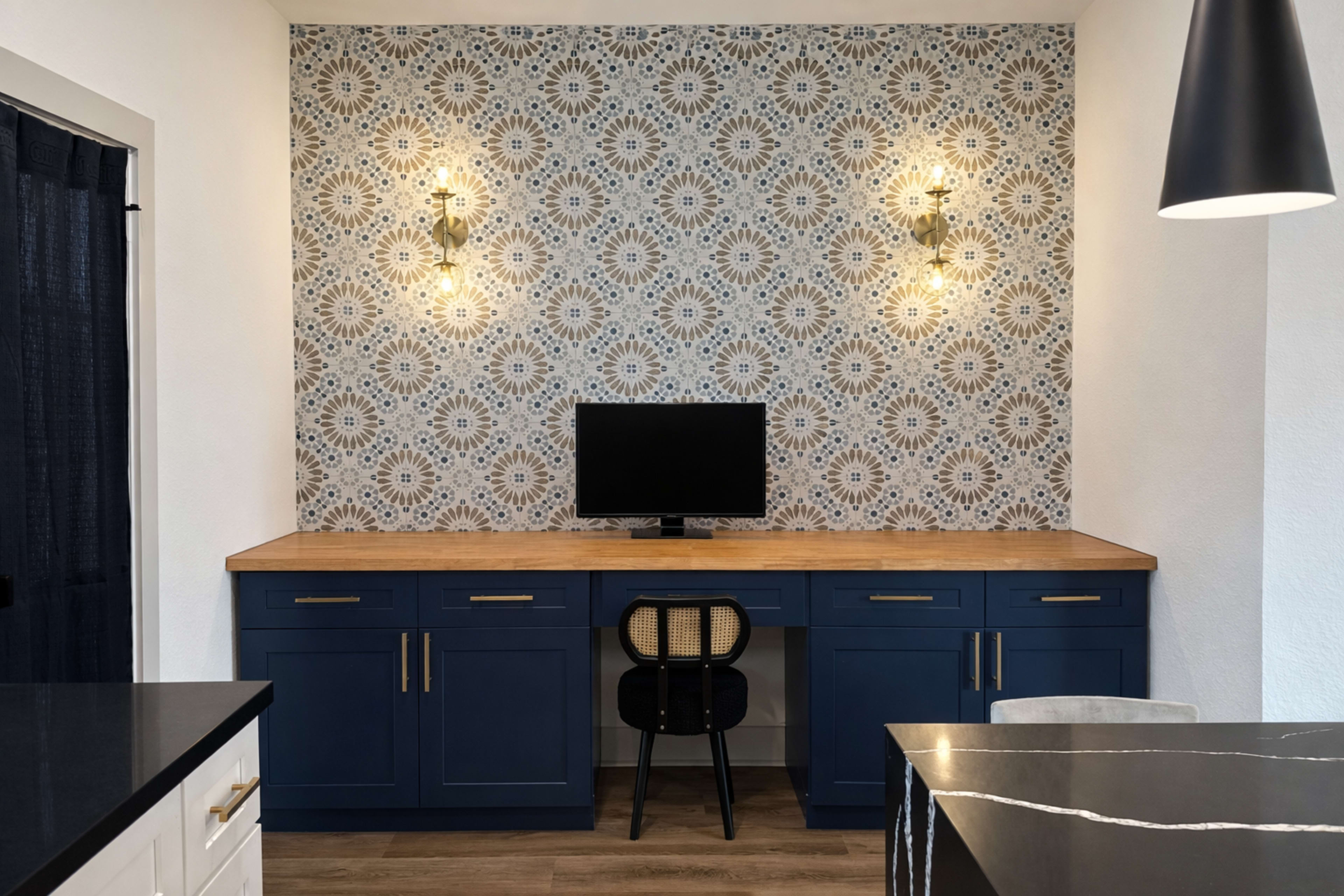 The image shows a modern workspace featuring a wooden desk with a black chair, a television on the wall, and decorative wallpaper with a floral pattern.