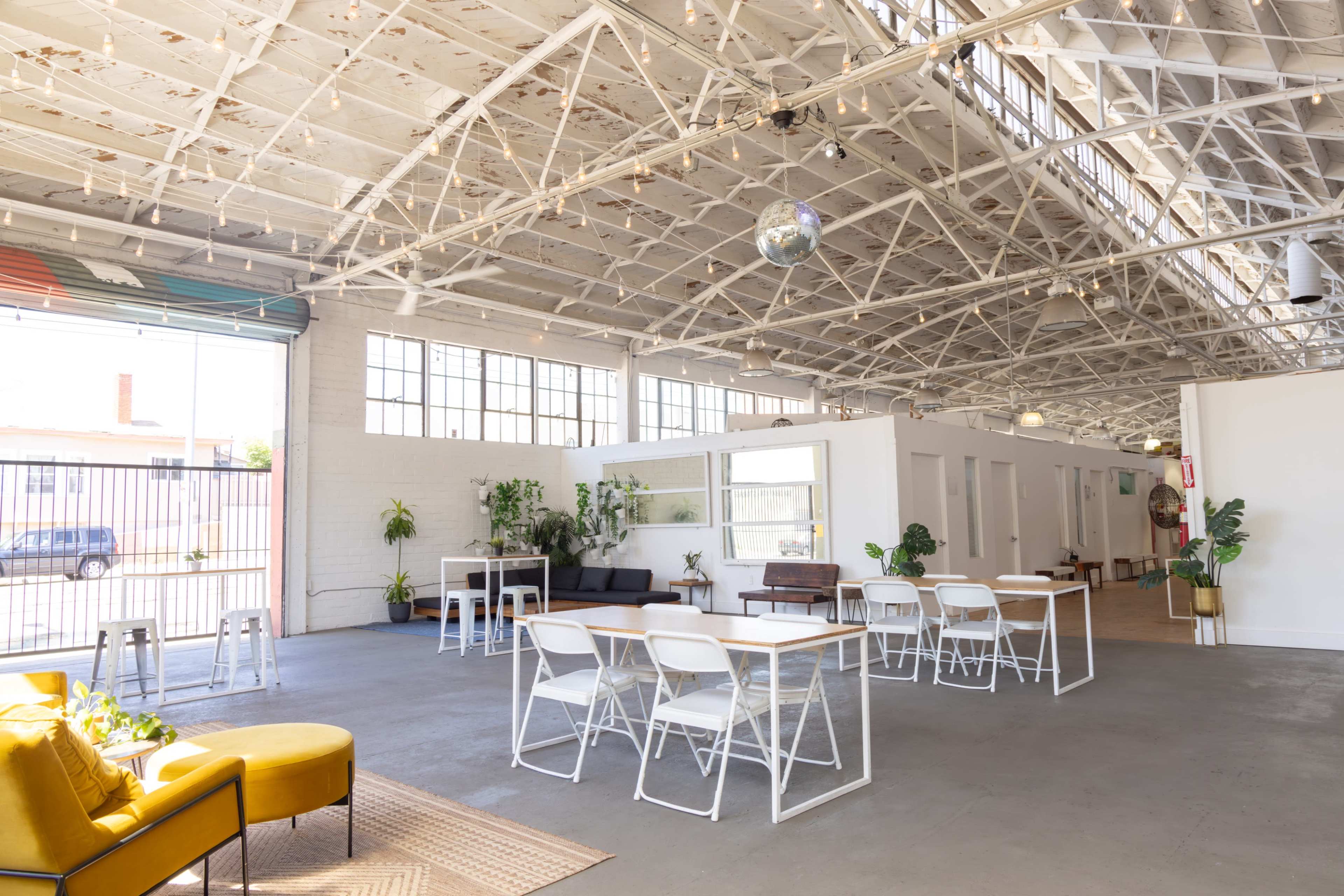 The image shows a spacious, industrial-style interior with a high ceiling, large windows, lounge seating, and a mix of dining and casual tables.