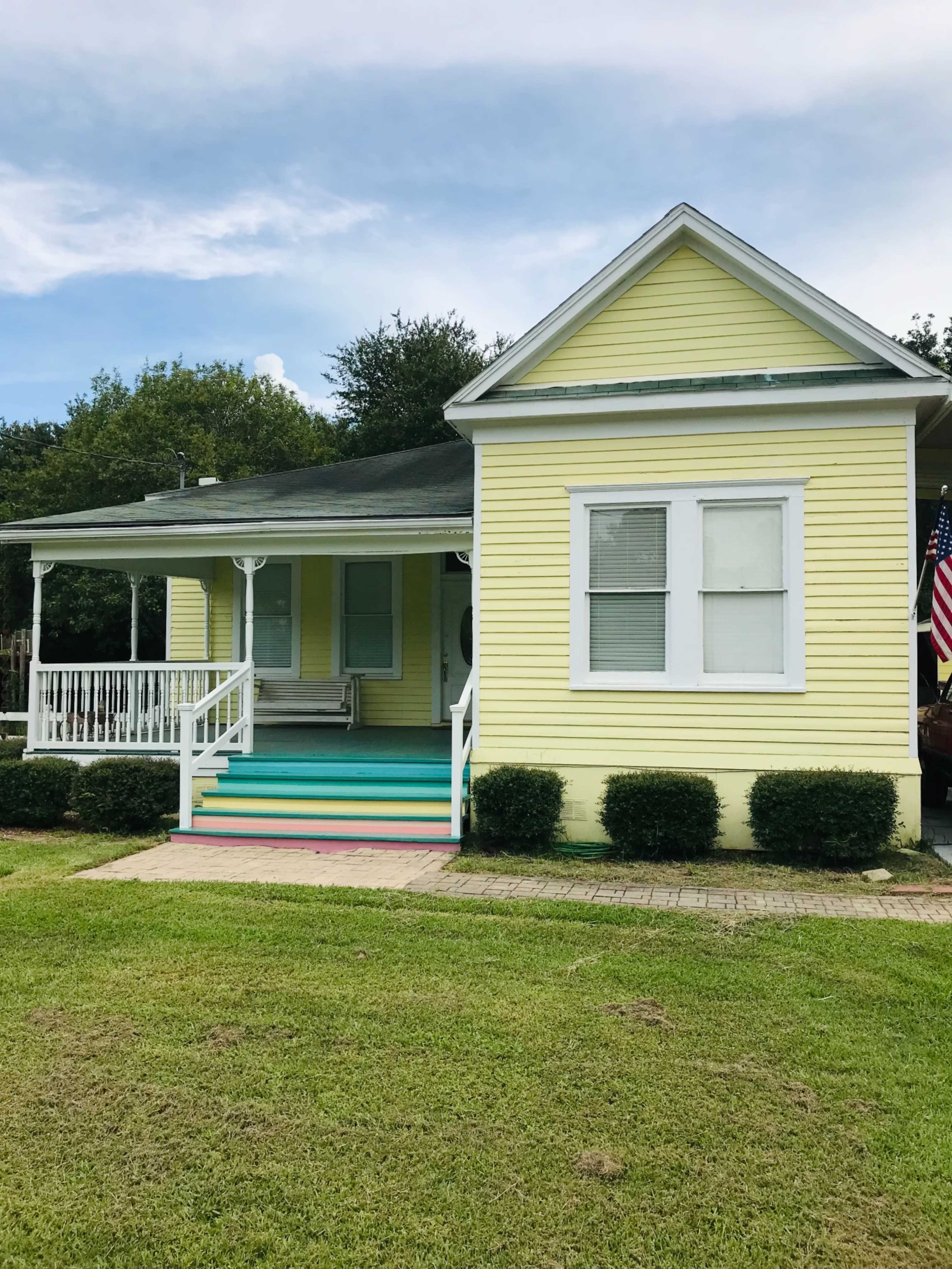 A yellow house with a covered porch and colorful steps is situated on a grassy lawn.