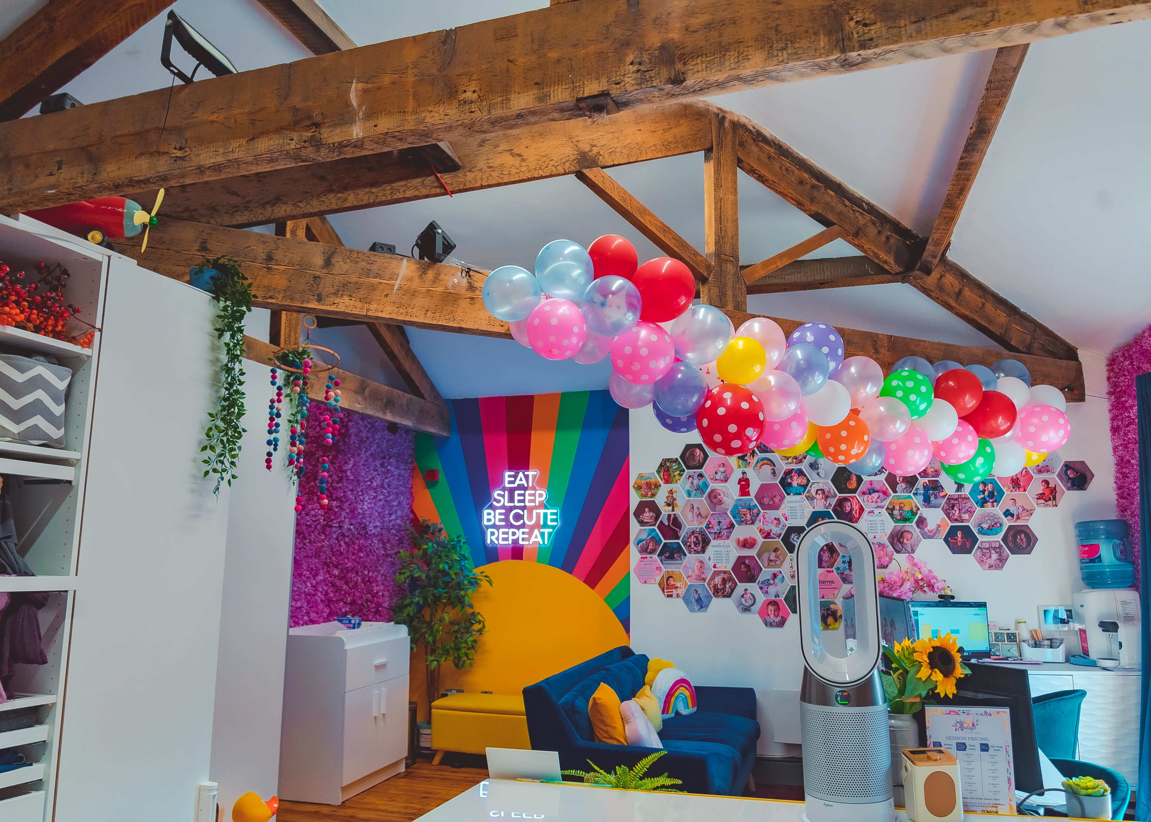 A brightly colored room features exposed wooden beams, a wall adorned with balloons and photographs, and a neon sign that reads "EAT SLEEP BE CUTE REPEAT."