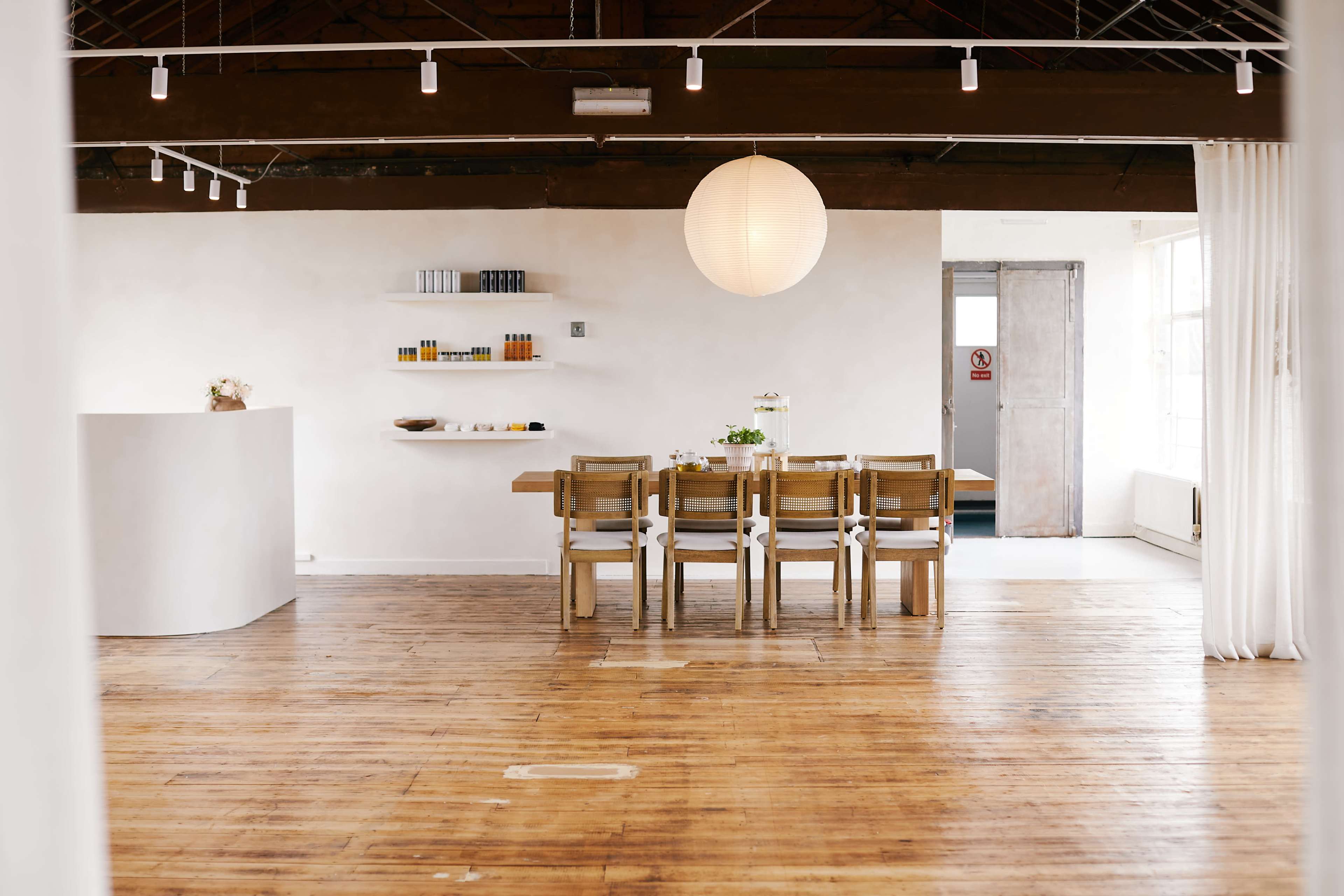 The image shows a spacious interior featuring a long wooden dining table surrounded by chairs, with a circular light fixture overhead and minimalist decor throughout.