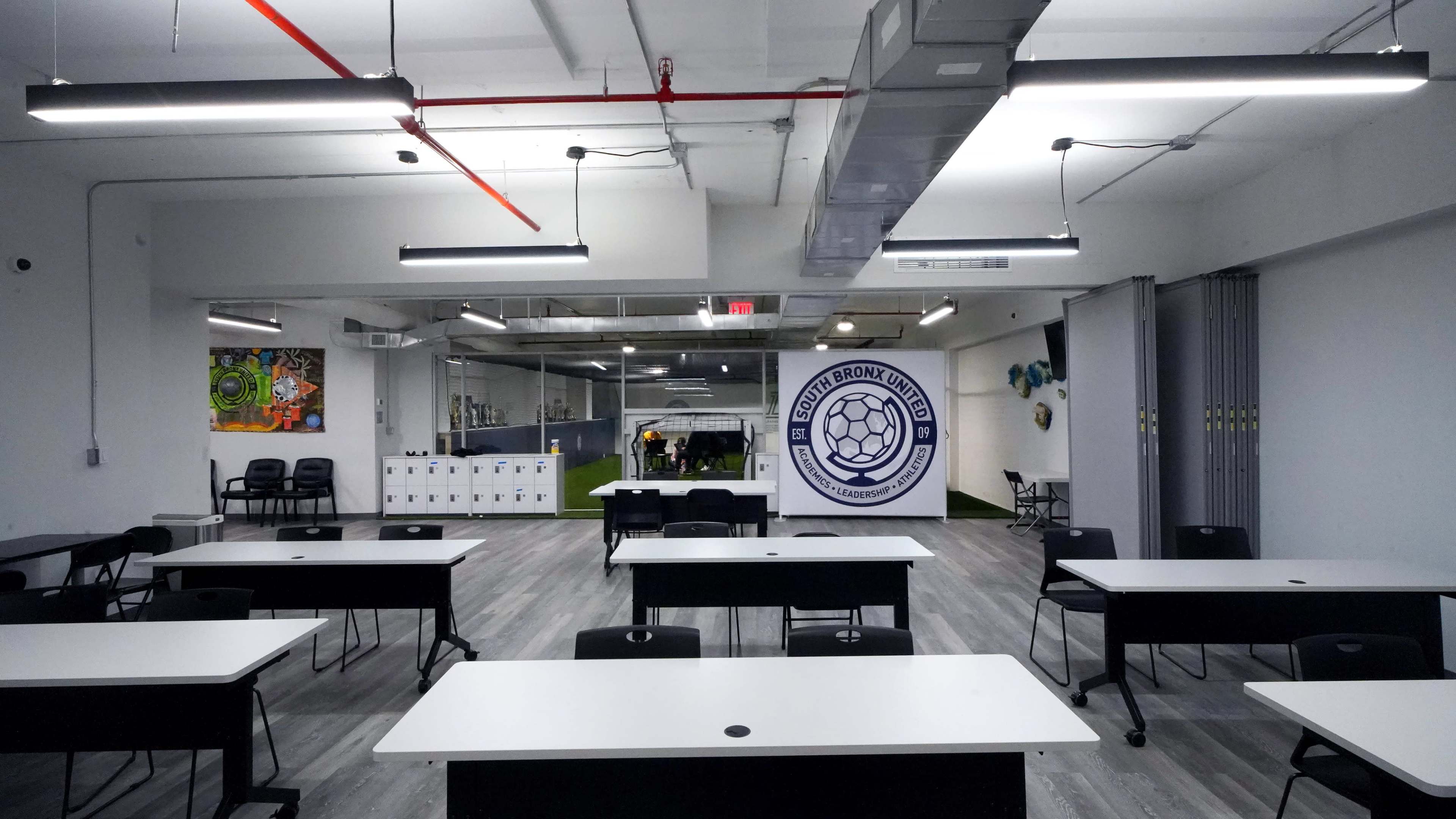 The image shows a modern classroom or meeting space with multiple tables and chairs arranged in rows, featuring a large logo of a soccer team on the wall.