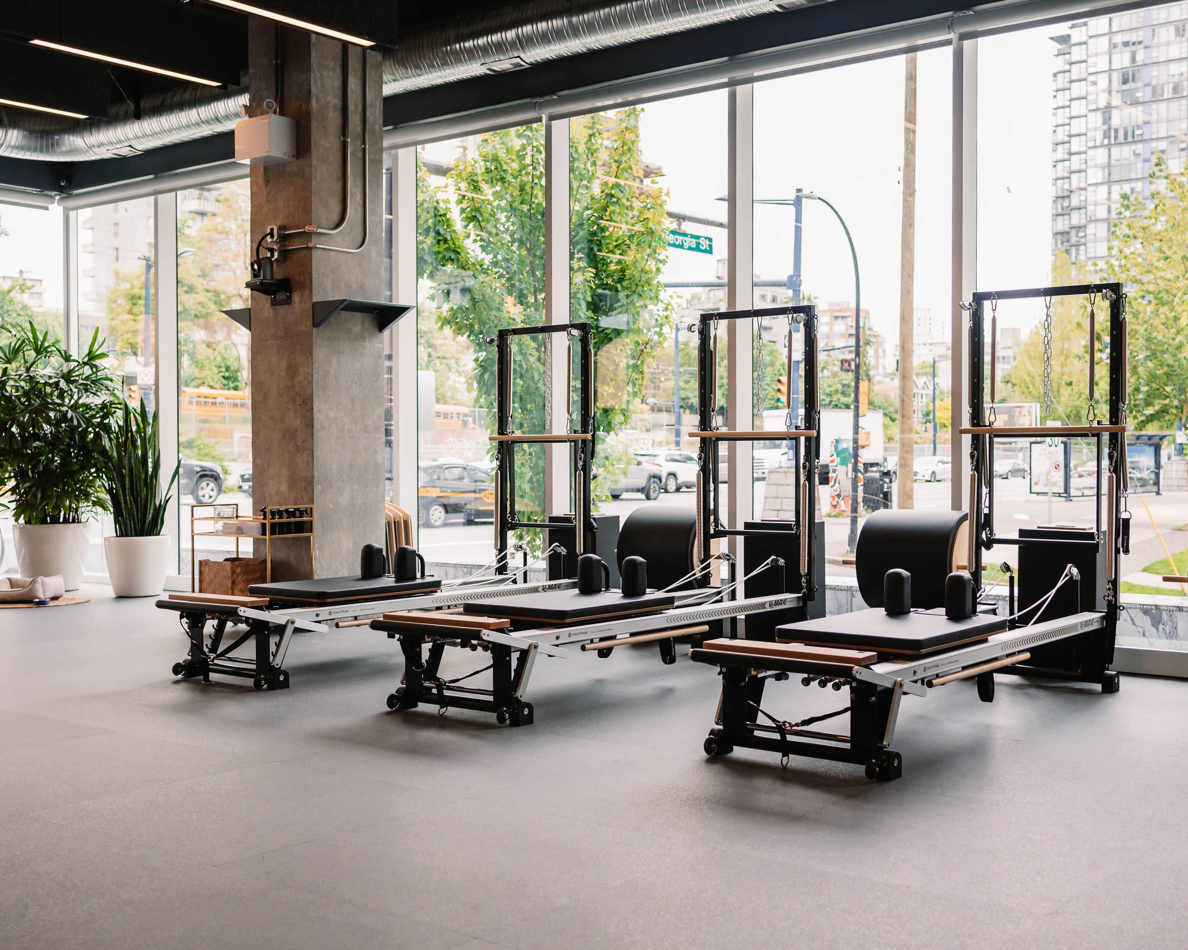 The image shows a bright fitness studio featuring several reformer Pilates machines set against large windows.