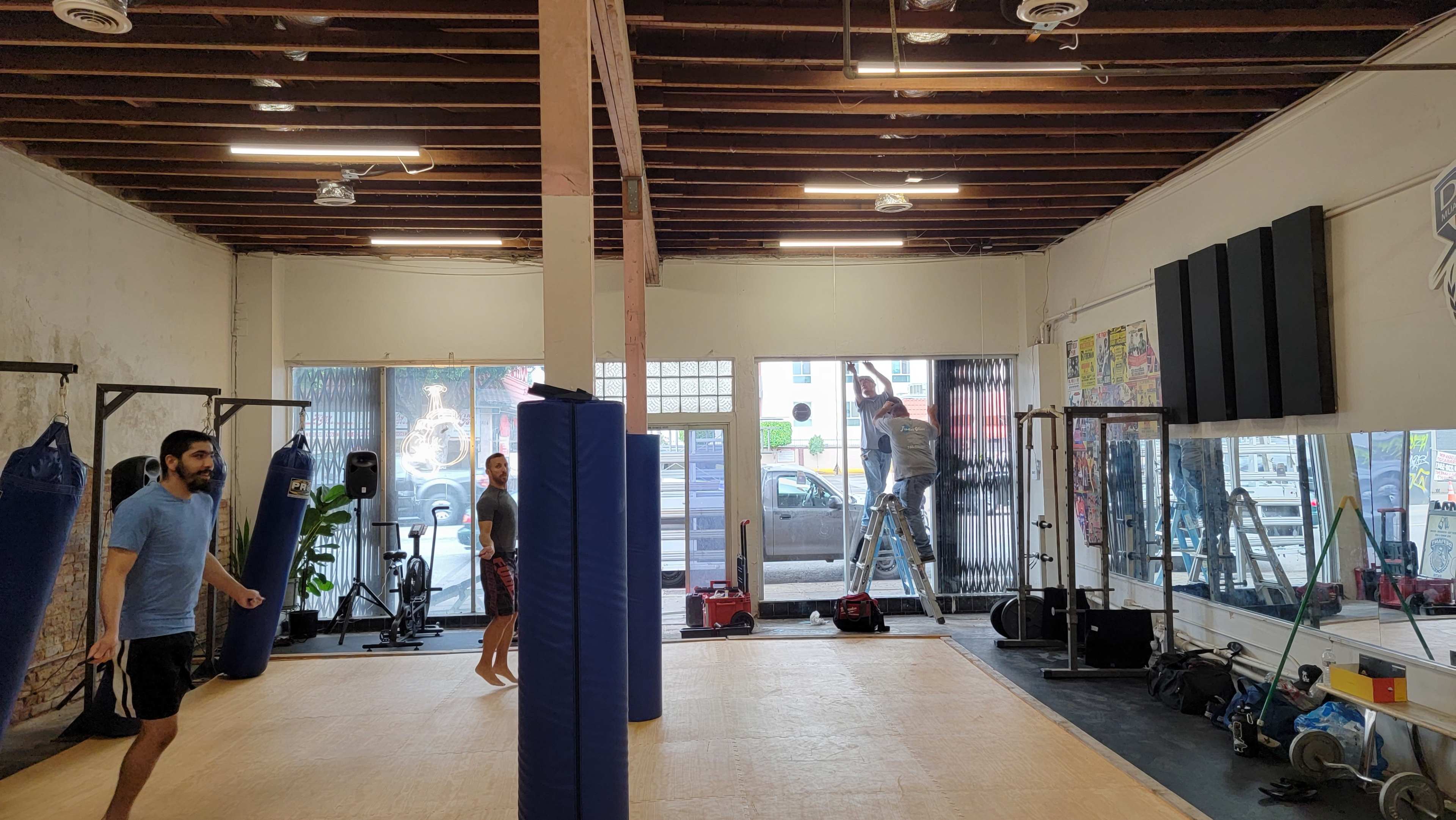 An indoor gym with a wooden floor, boxing bags on the walls, and people training while construction work is happening in the background.