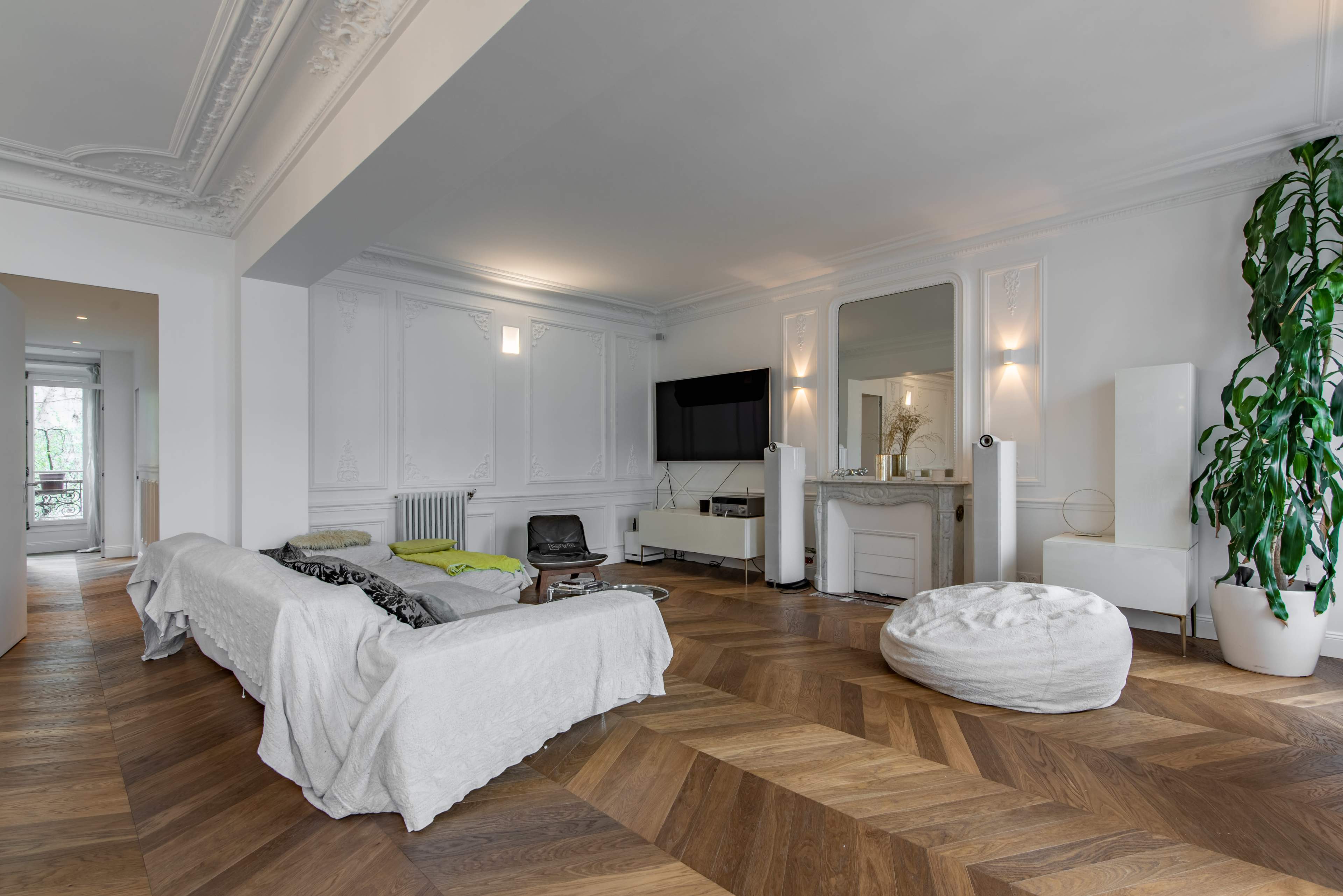 The room features a minimalist design with a sofa draped in a white cover, a television mounted on the wall, and a large plant beside a seating area on hardwood flooring.