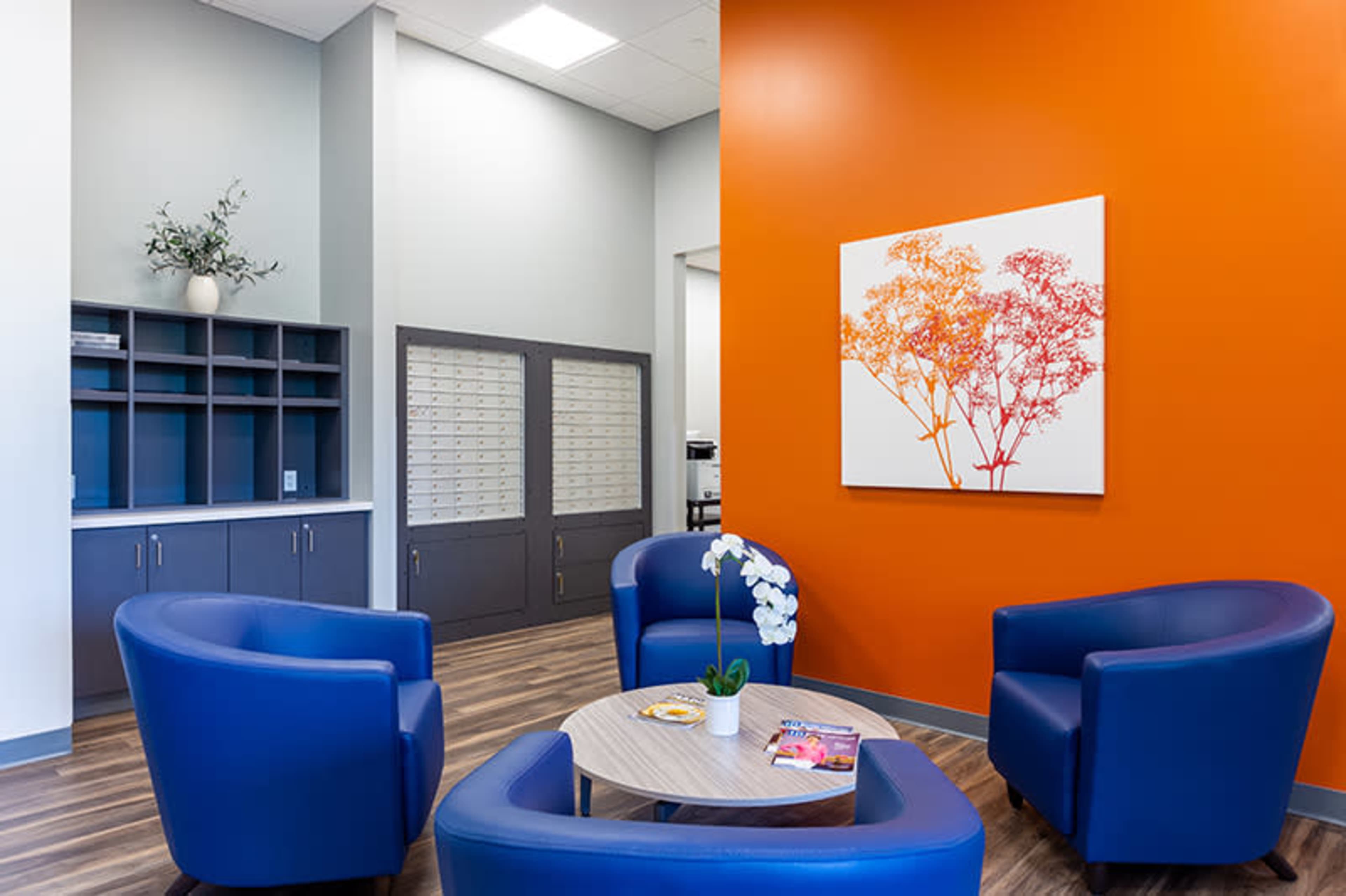 A modern waiting area features blue chairs arranged around a circular table, with an orange accent wall and a floral artwork.