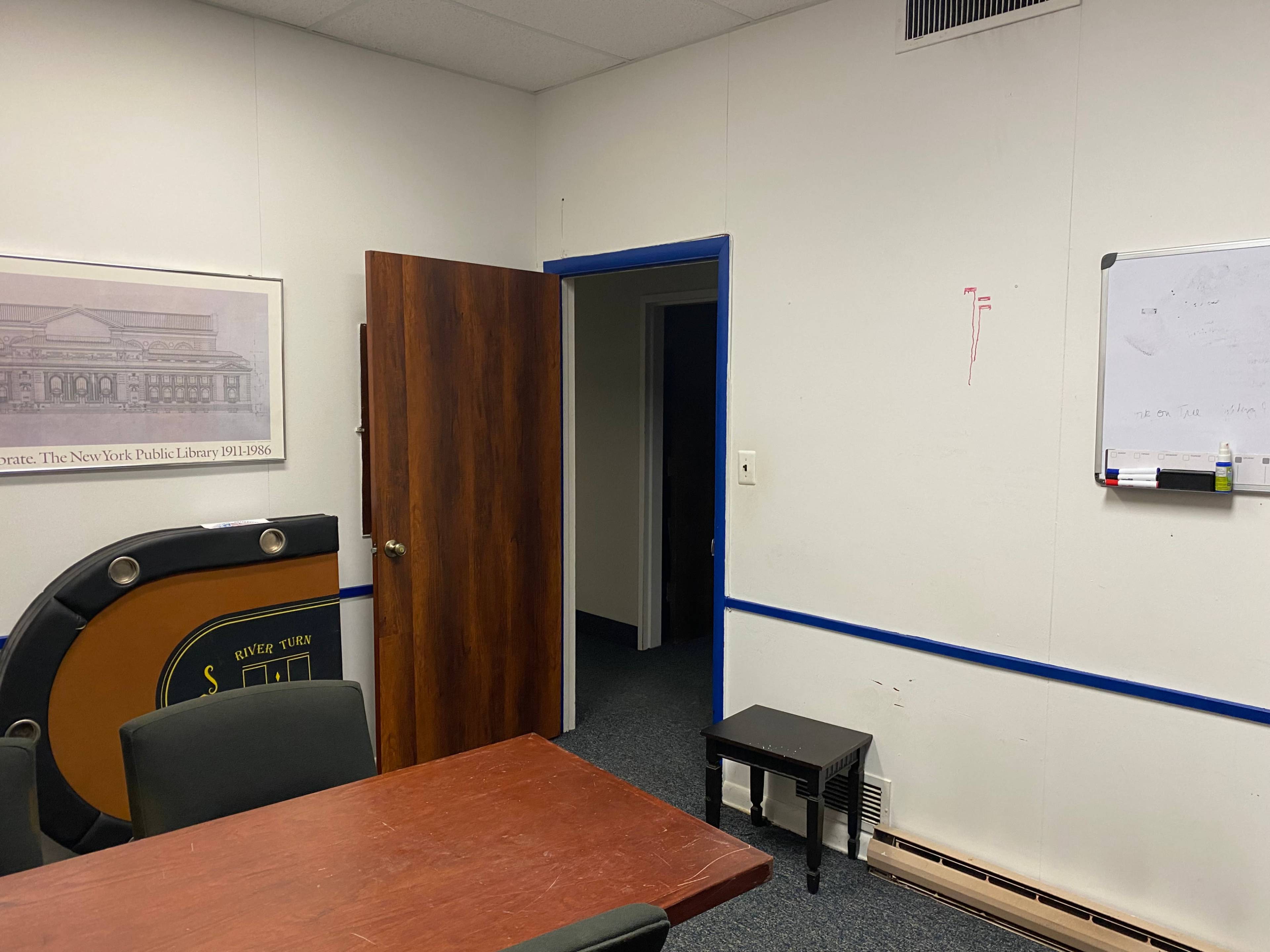 The image shows a small office space with a wooden table, chairs, a door leading to another room, and a whiteboard on the wall.