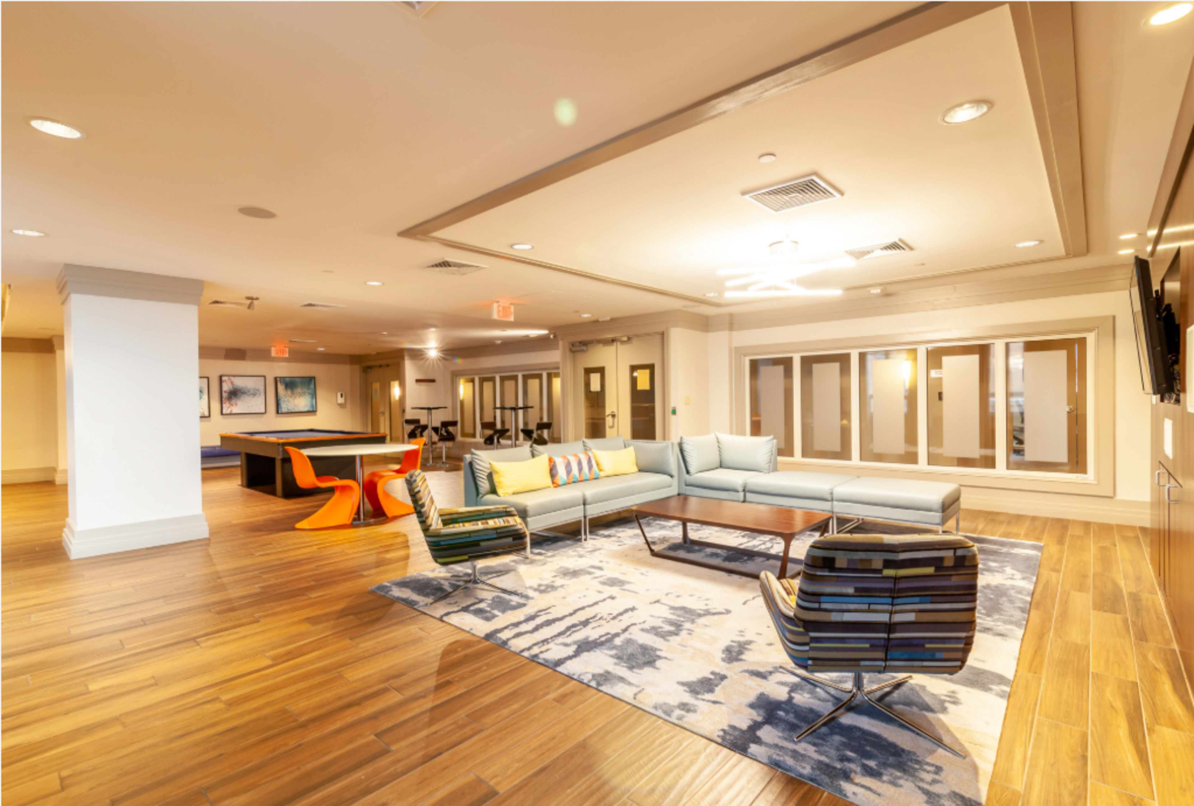 The image shows a modern lounge area featuring a seating arrangement with colorful chairs and a large rug, alongside a pool table and sliding doors.