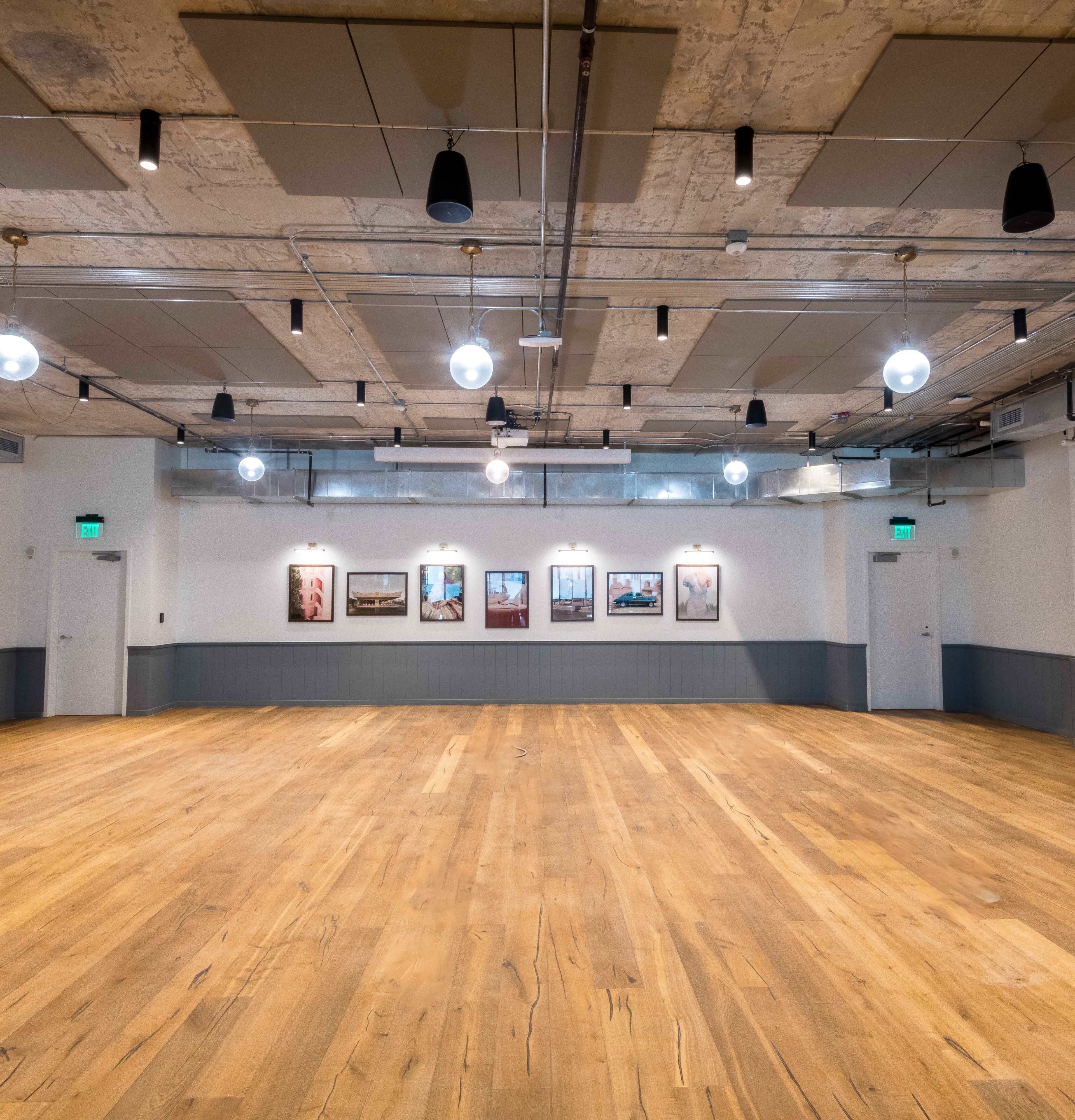 A spacious, empty room with wooden flooring, industrial lighting, and framed artwork on the walls.