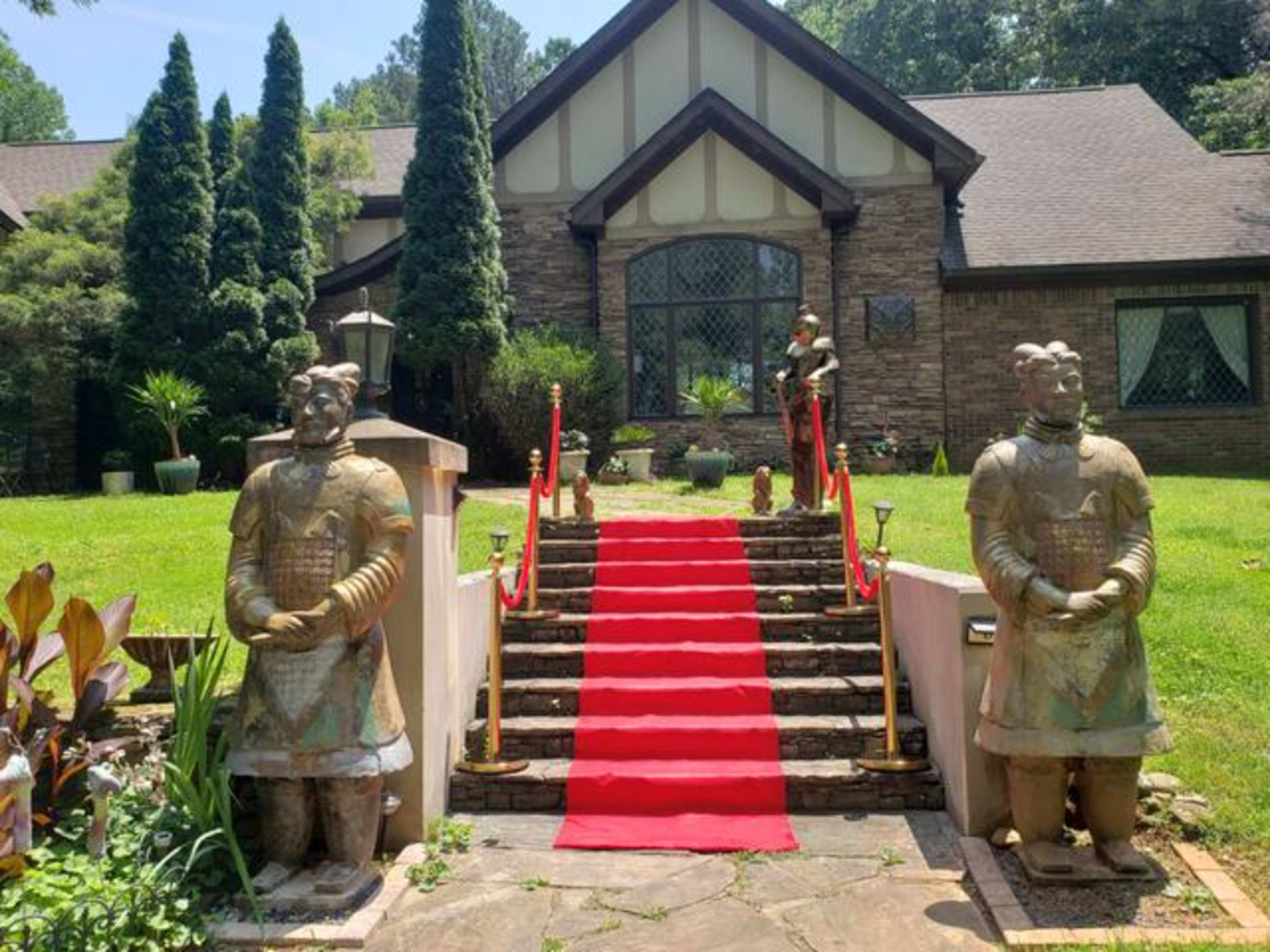 A house with a stone facade and a red carpet leading up to the entrance, flanked by two large statues resembling ancient warriors.