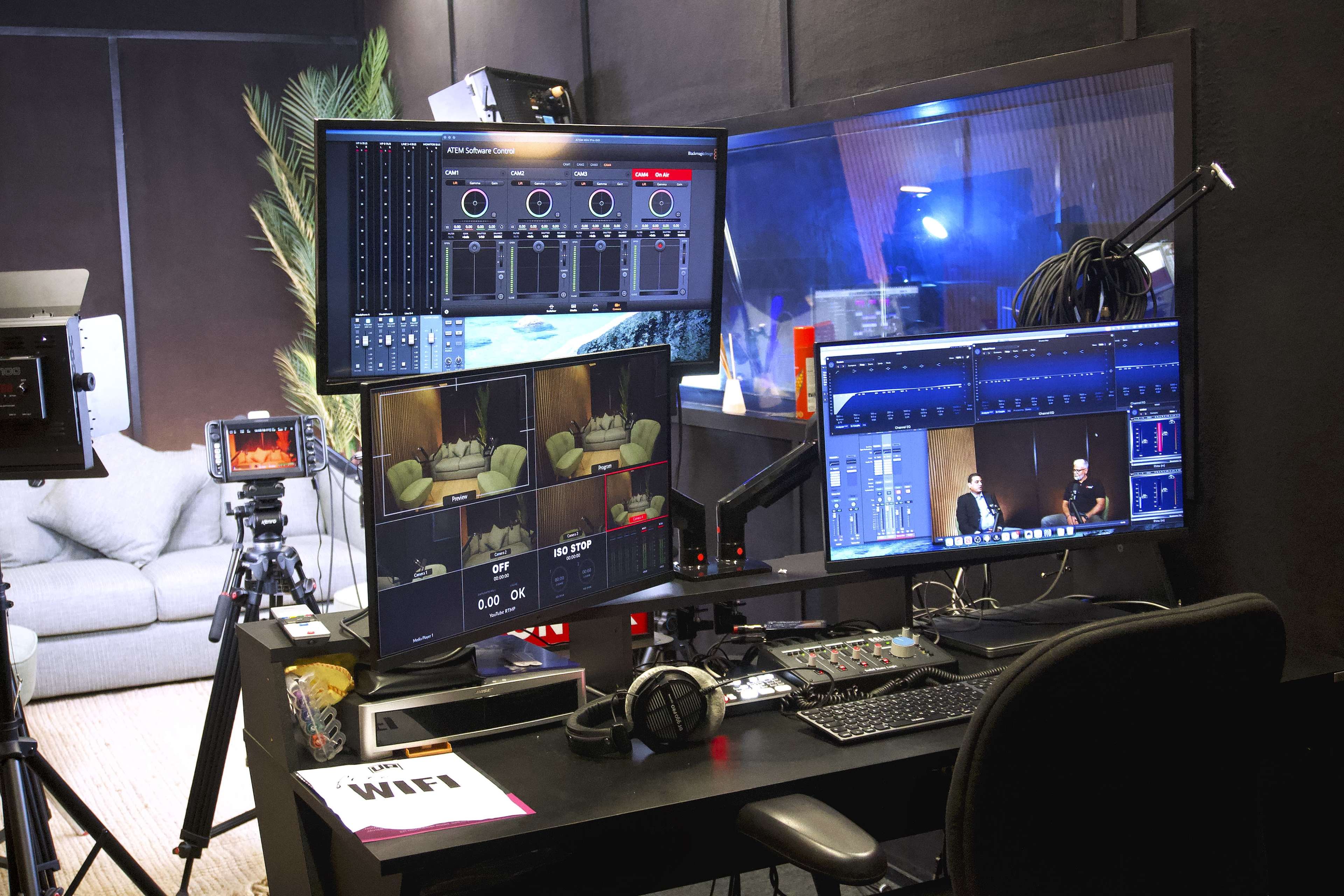 The image shows a broadcast studio setup with multiple monitors displaying audio and video controls, a camera, and various production equipment arranged on a black desk.