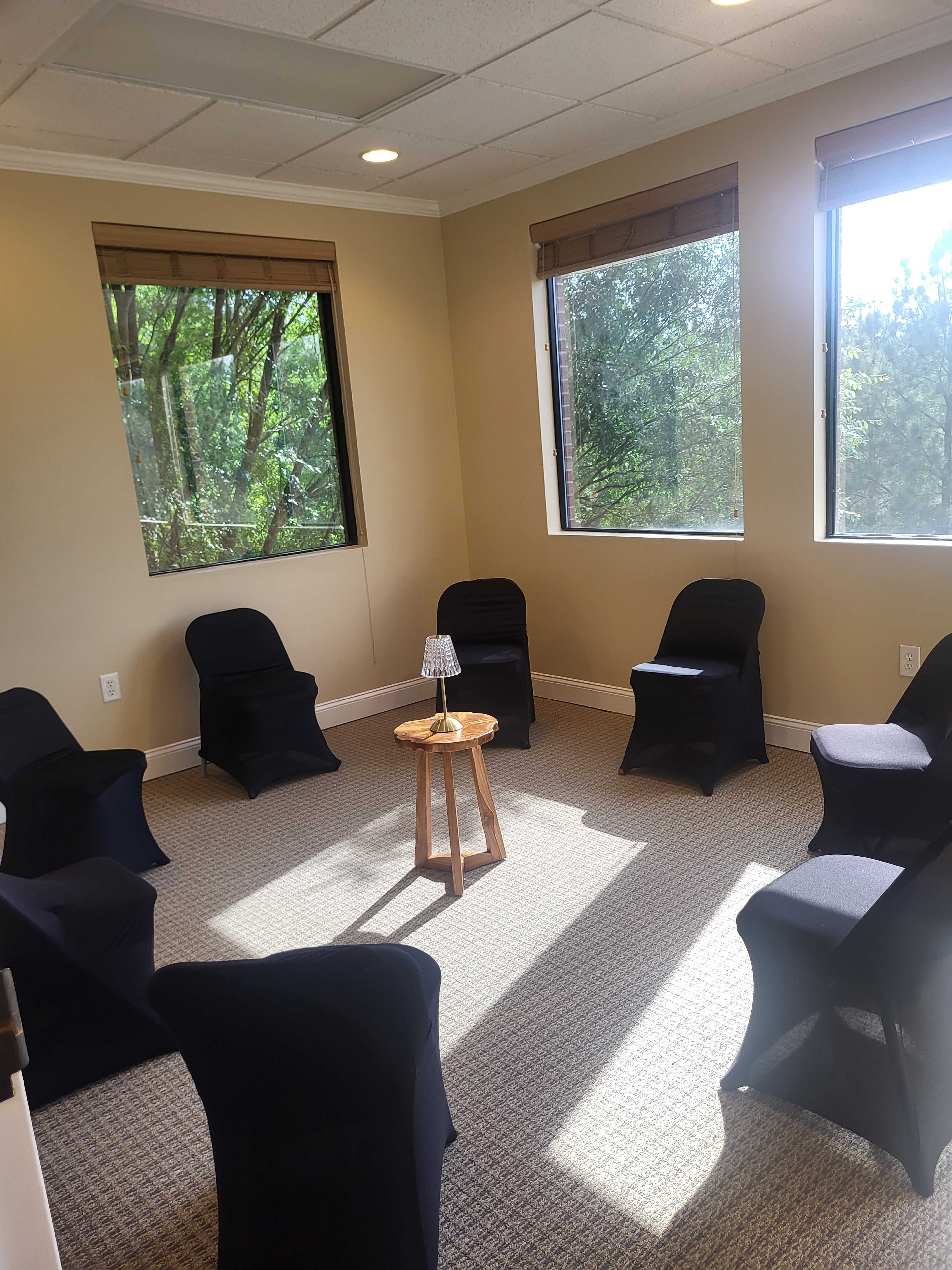 The image shows a small, well-lit meeting room with six dark chairs arranged in a circle around a small wooden table with a lamp.