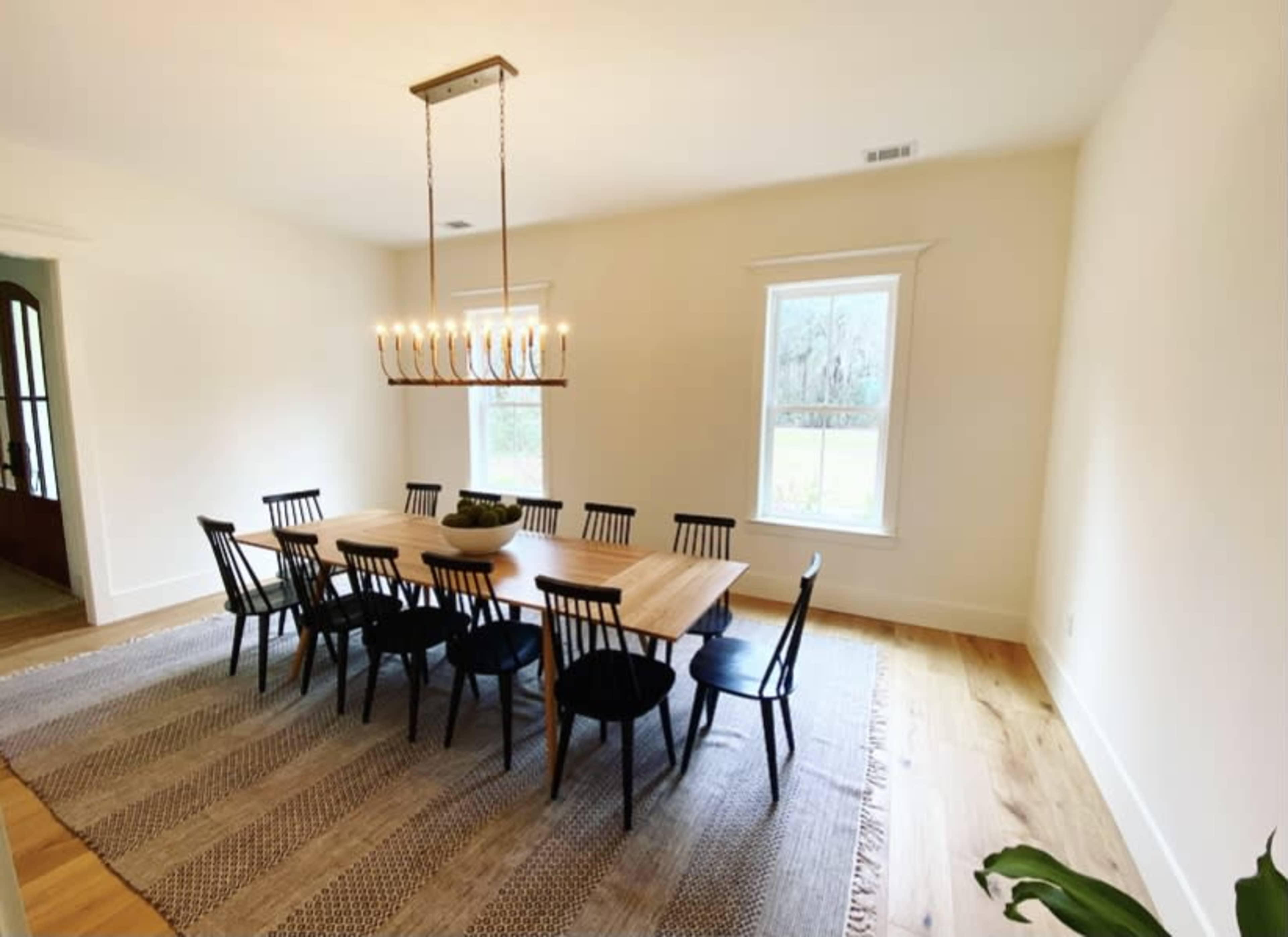 A long wooden dining table with black chairs is located in a light-filled room with two windows and a chandelier overhead.