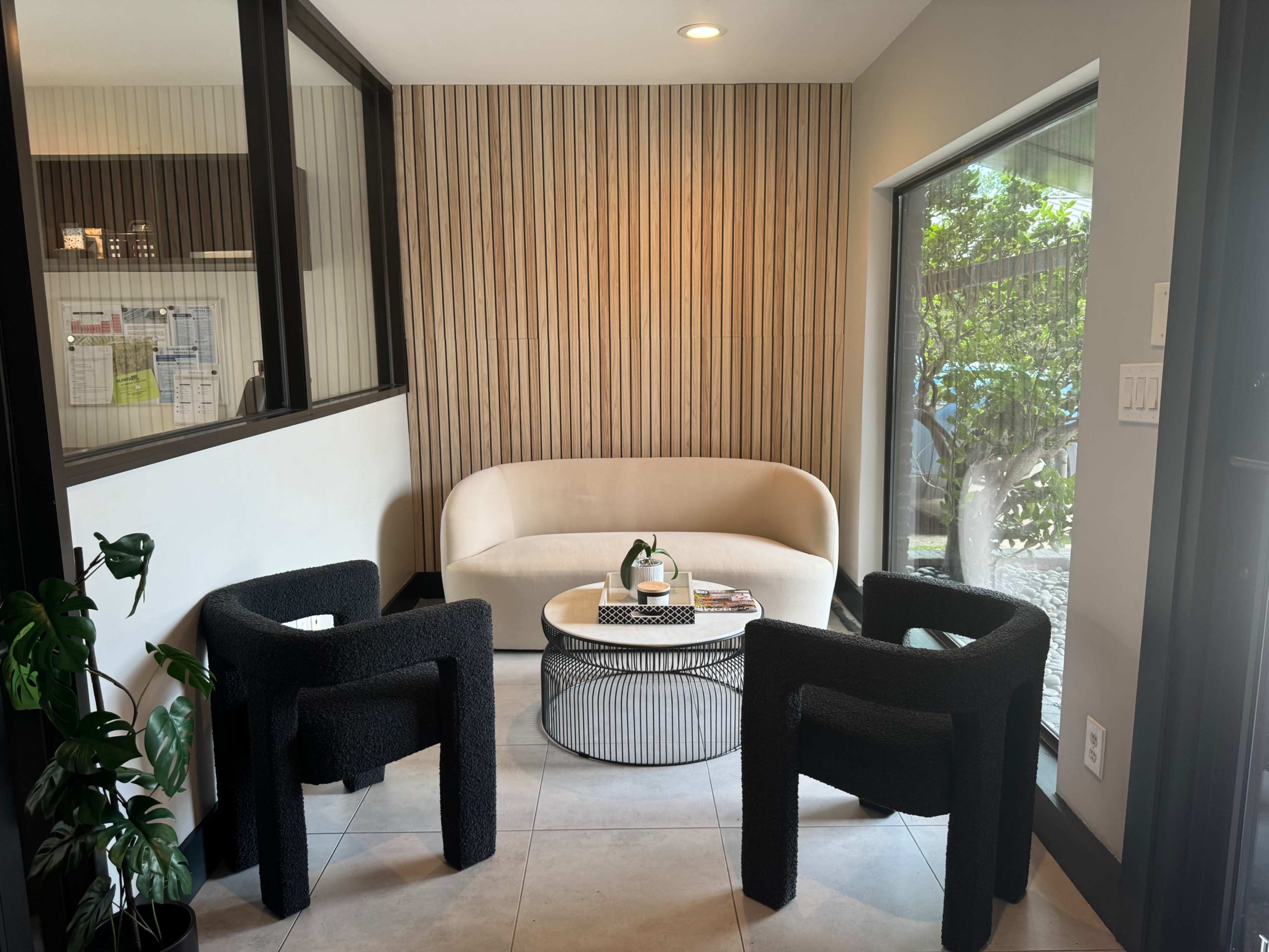 The image shows a modern sitting area with a curved sofa, two textured black chairs, and a round coffee table, all situated near large windows letting in natural light.