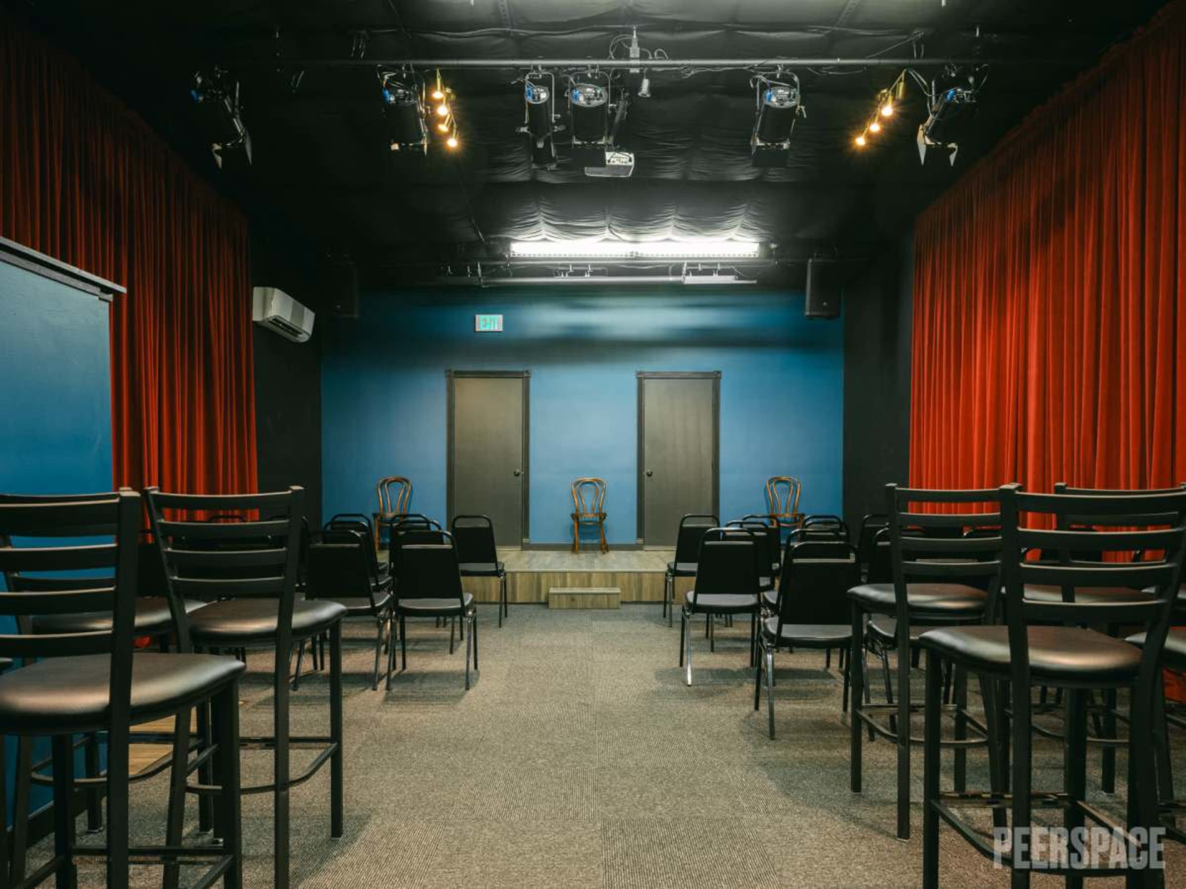 A small theater space with black chairs arranged facing a stage, flanked by red curtains and two closed doors in the back.