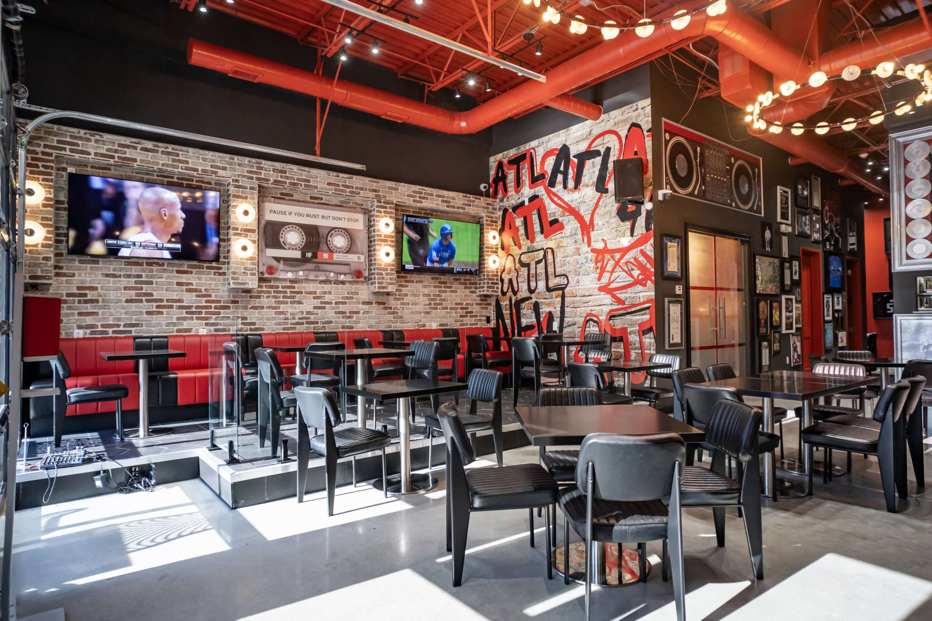 The image shows a modern restaurant interior with exposed brick walls, red accents, and various seating arrangements, including tables and bar-style seating, complemented by multiple televisions mounted on the walls.