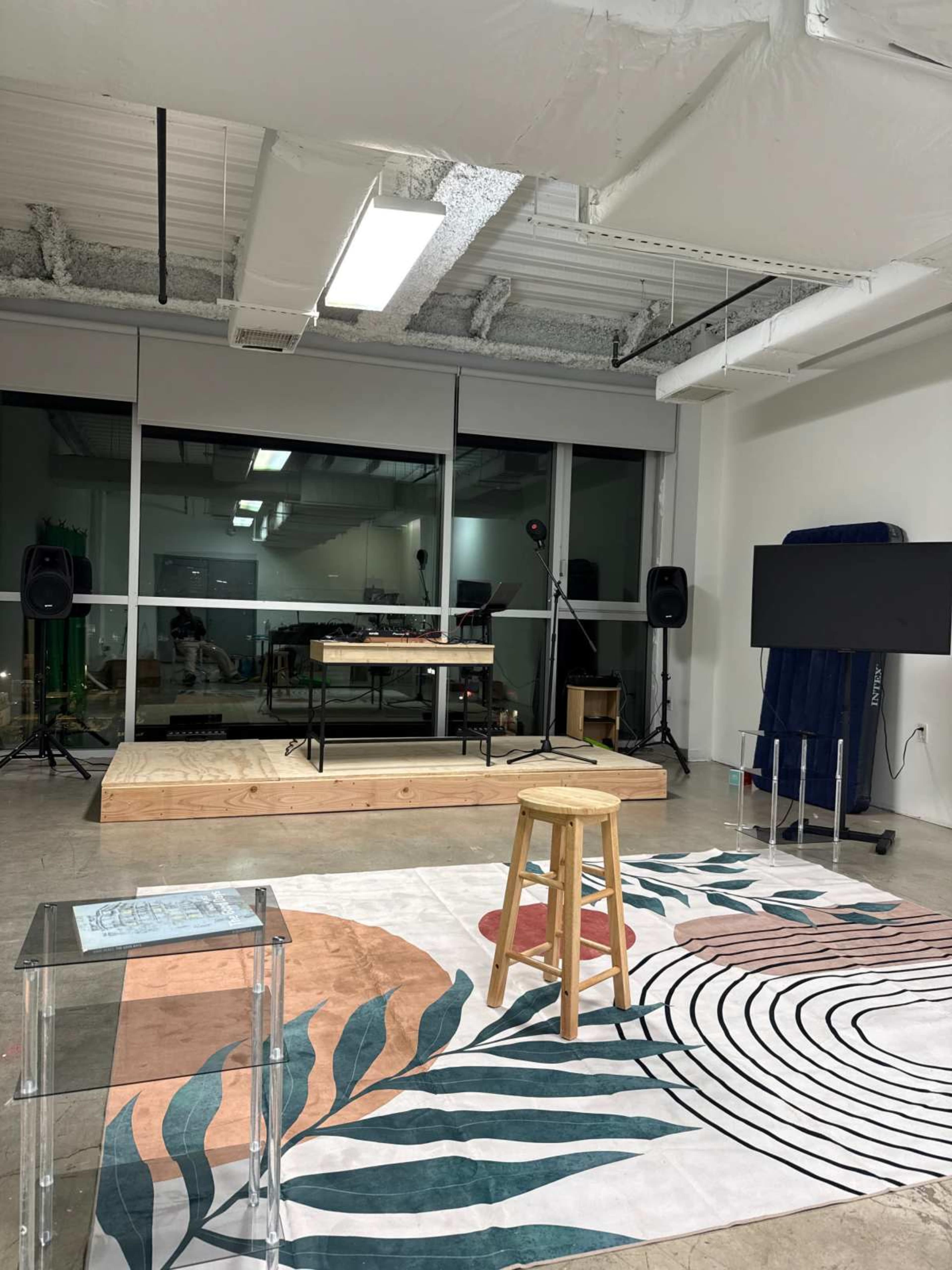 The image shows a modern studio space with a large patterned rug, a wooden stage, a stool, and audio equipment set up on either side, along with a large screen on a stand.