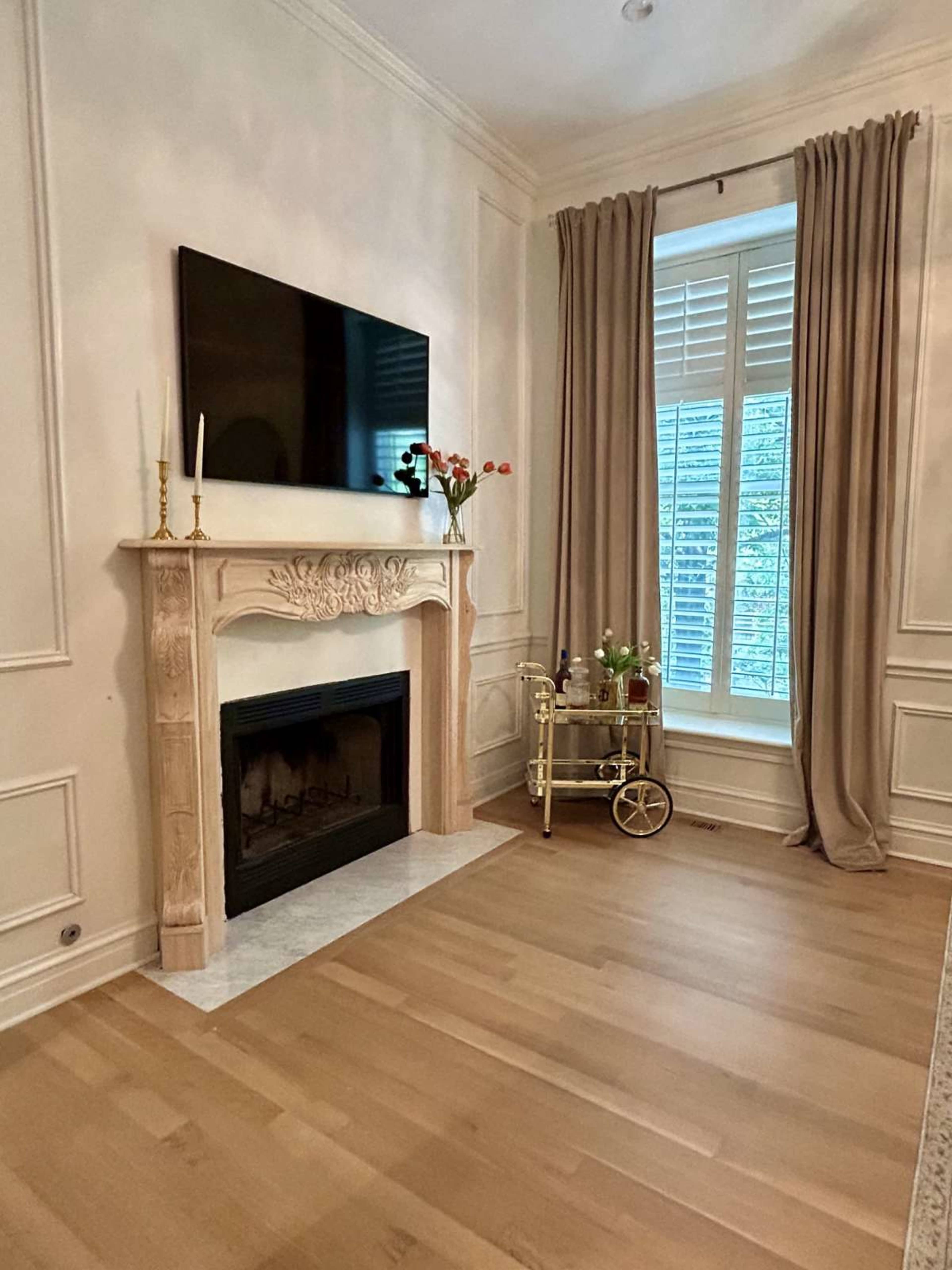 The image shows a living room featuring a fireplace with a TV above it, a window with curtains, and a small rolling cart beside the fireplace.