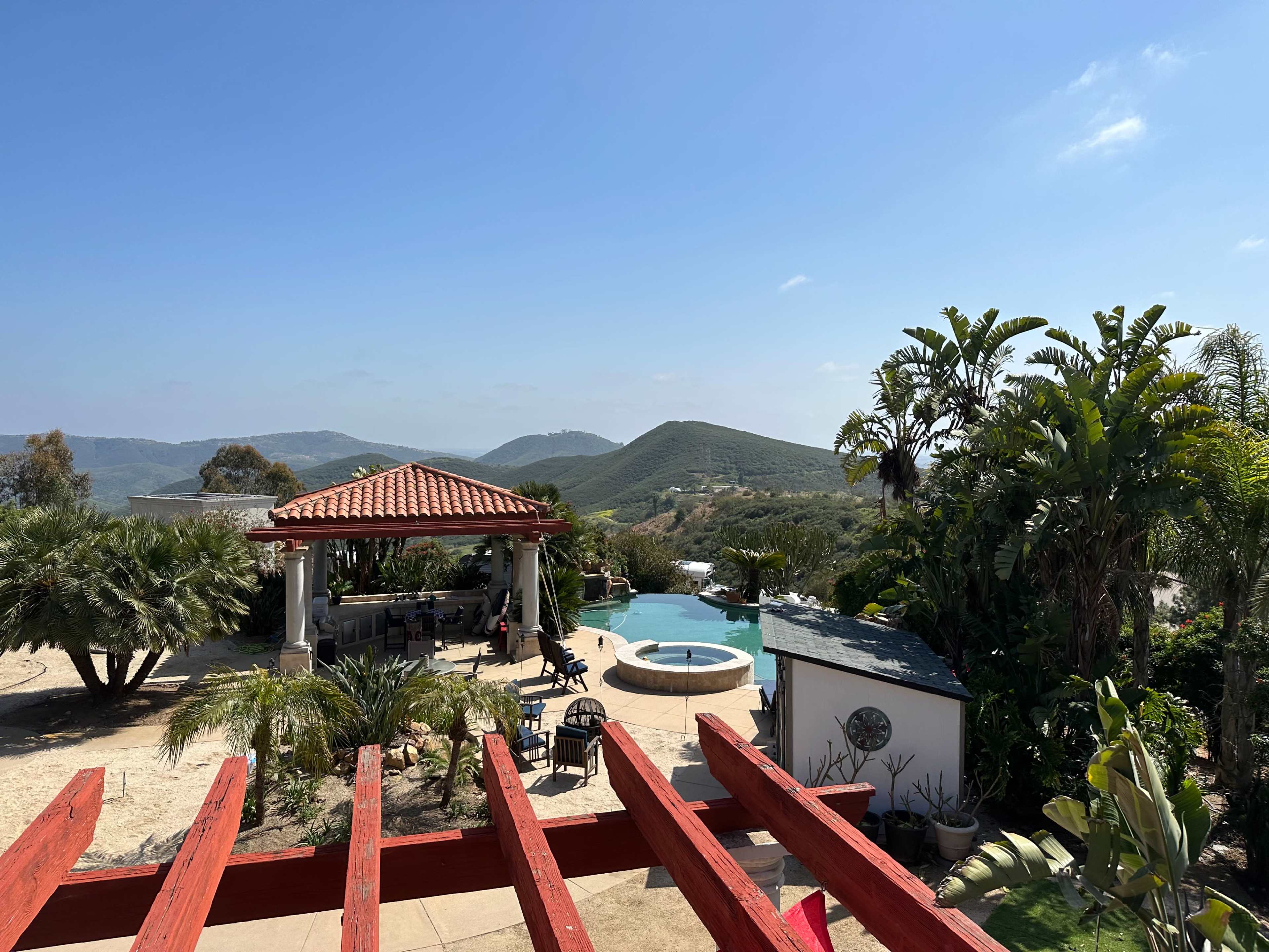 The image shows a patio area with a swimming pool, a jacuzzi, and a garden overlooking rolling hills under a clear blue sky.