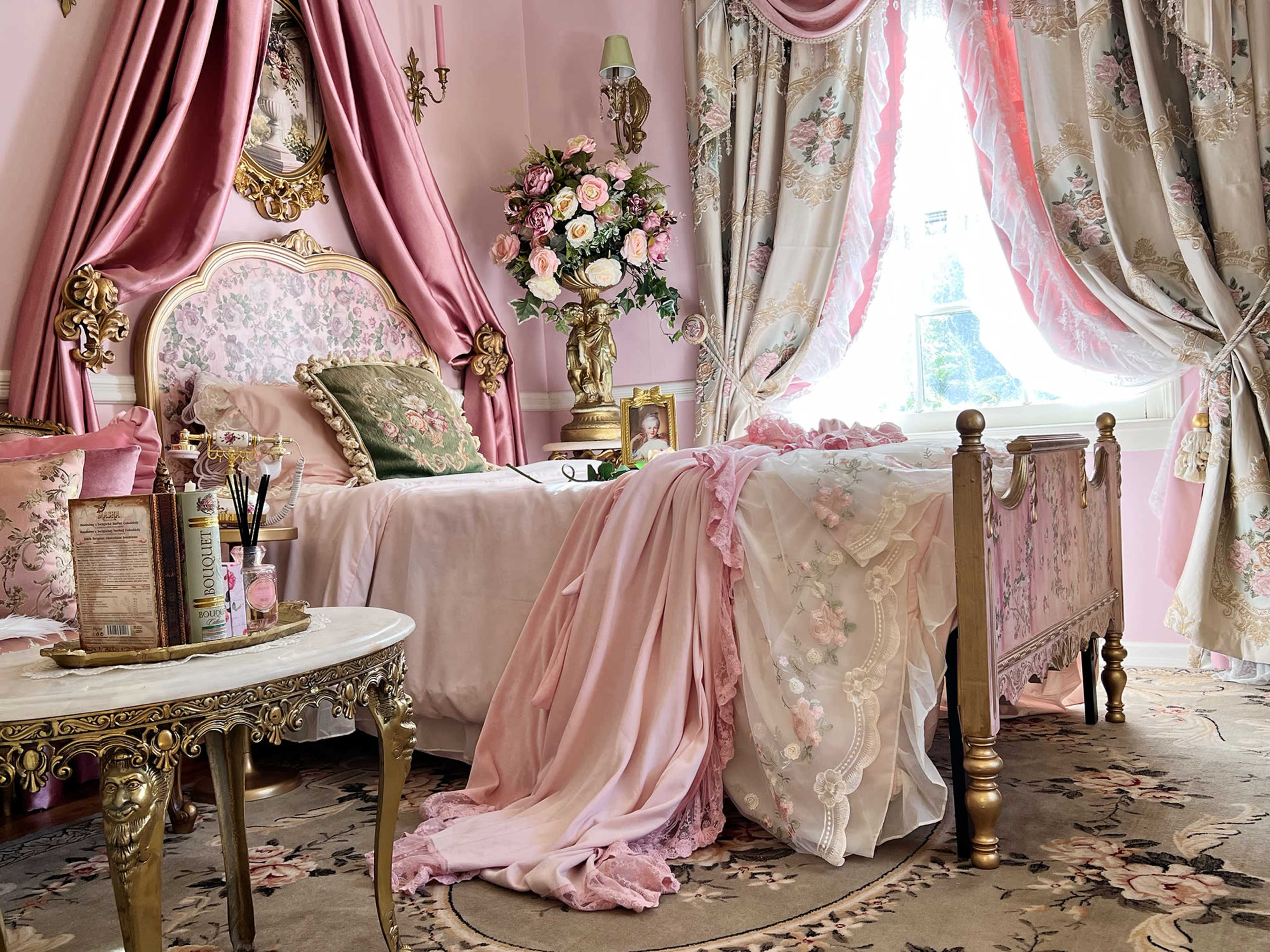 The image shows a feminine bedroom with a pink color scheme, featuring a floral-patterned bedspread, ornate furniture, and large draped curtains.