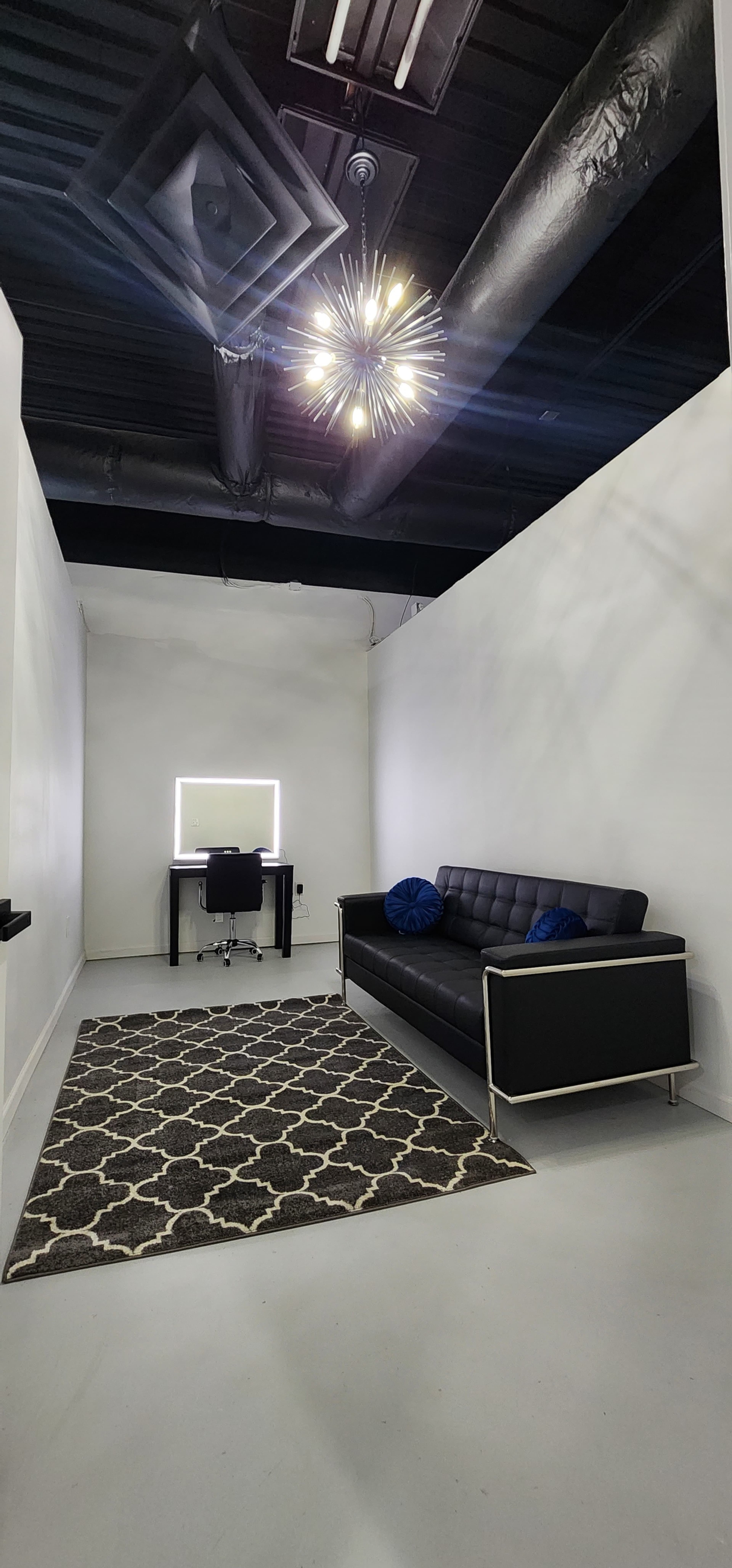 The image shows a minimalist office space featuring a black couch, a desk with a chair, and a patterned rug under ambient lighting.