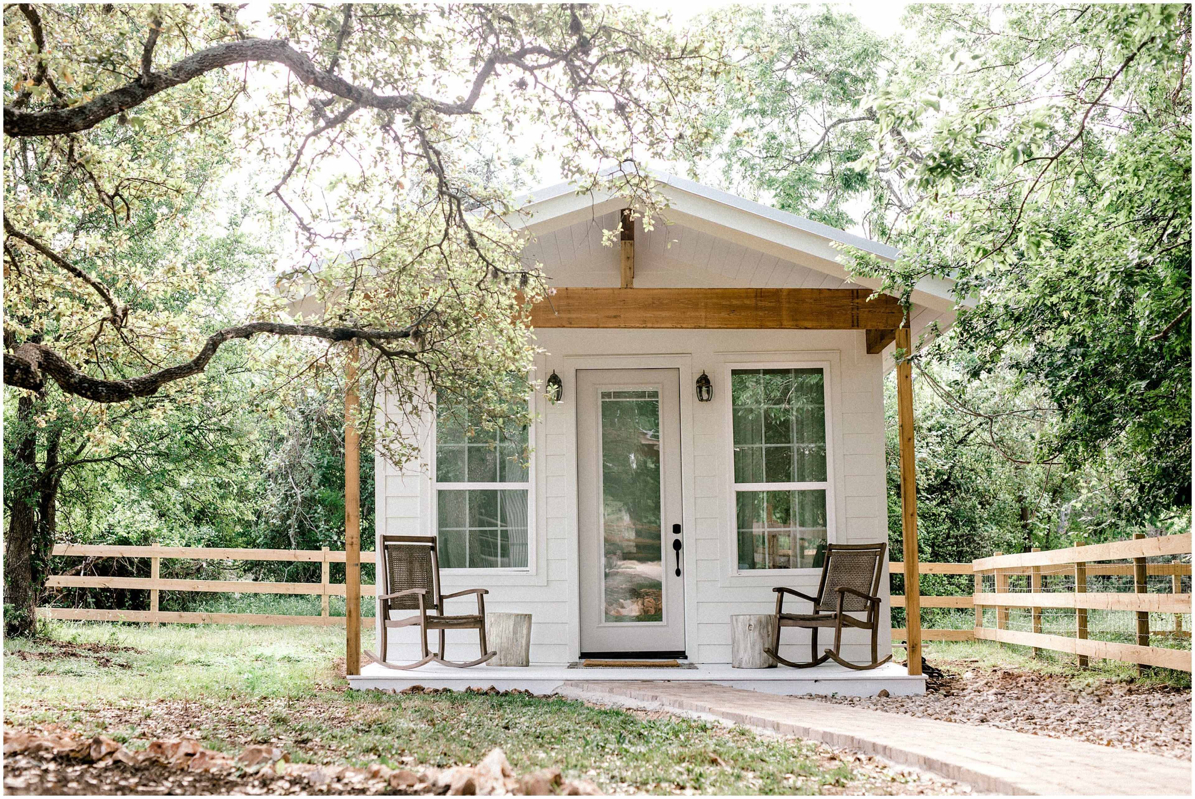 A small white cottage with a front porch and two wooden chairs is surrounded by trees and a wooden fence.