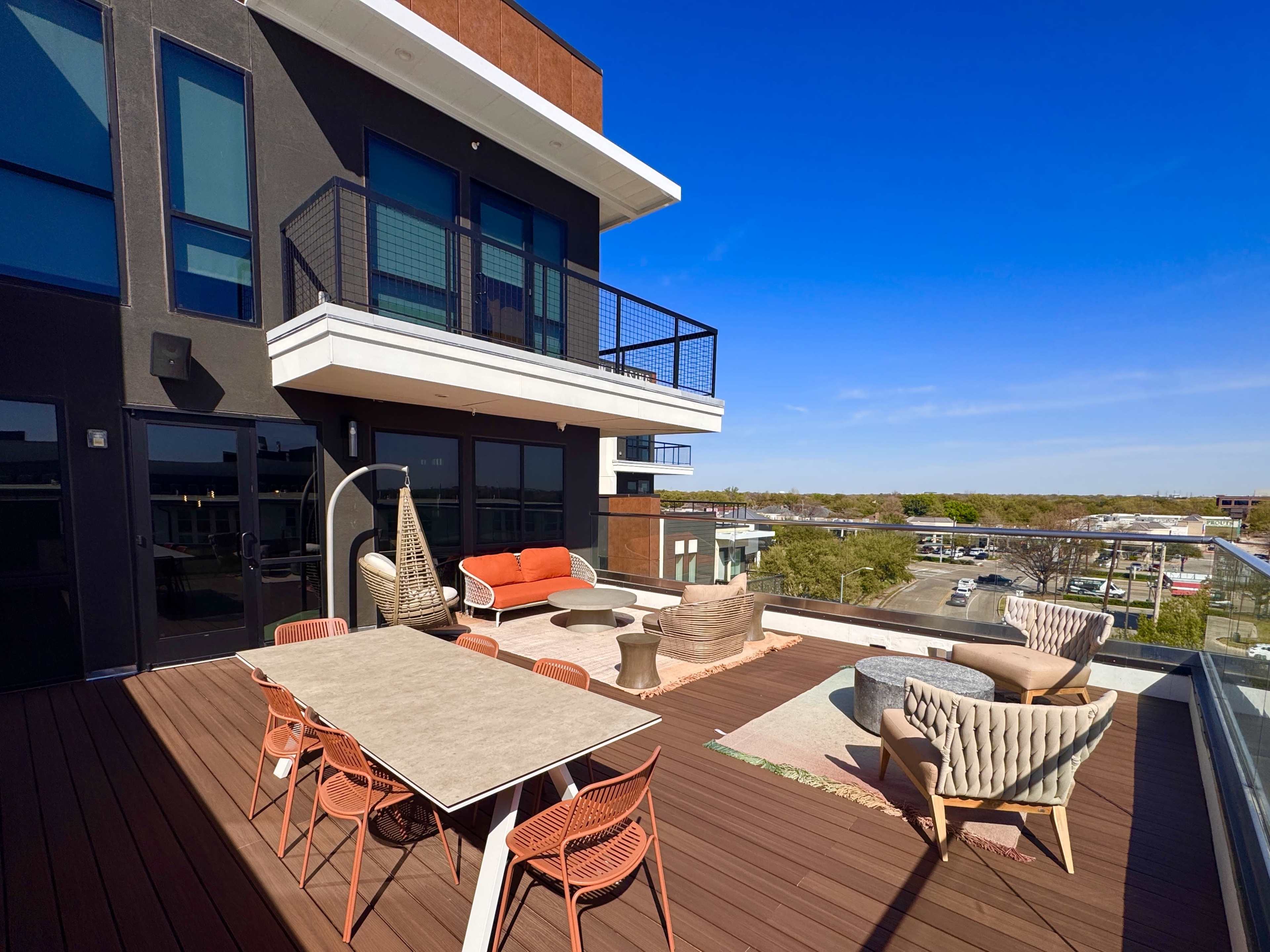 The image shows a modern rooftop terrace featuring a dining table, chairs, and various seating areas, surrounded by a clear blue sky.
