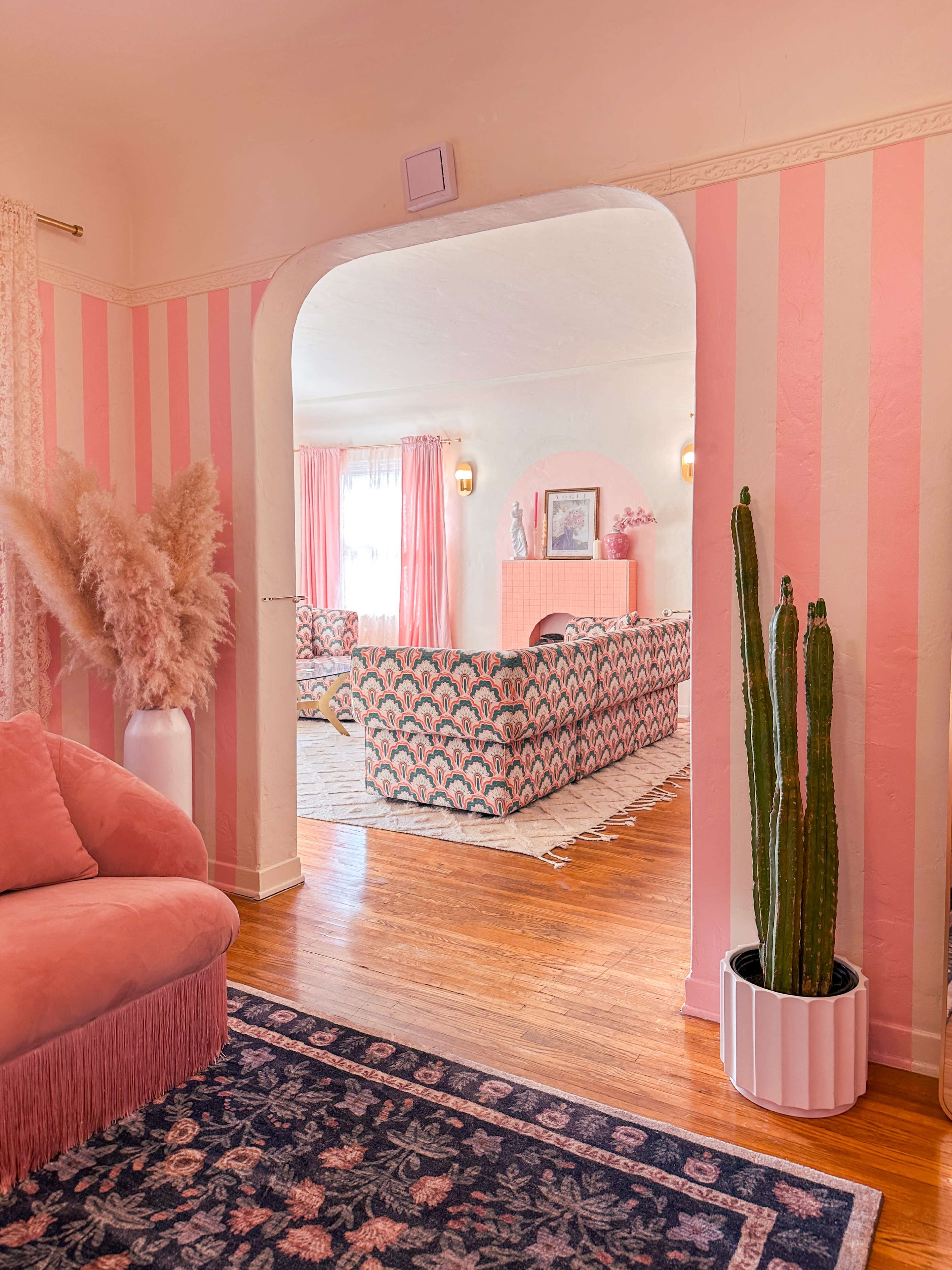 A room features pink striped walls, a patterned sofa, a potted cactus, and large windows allowing natural light.