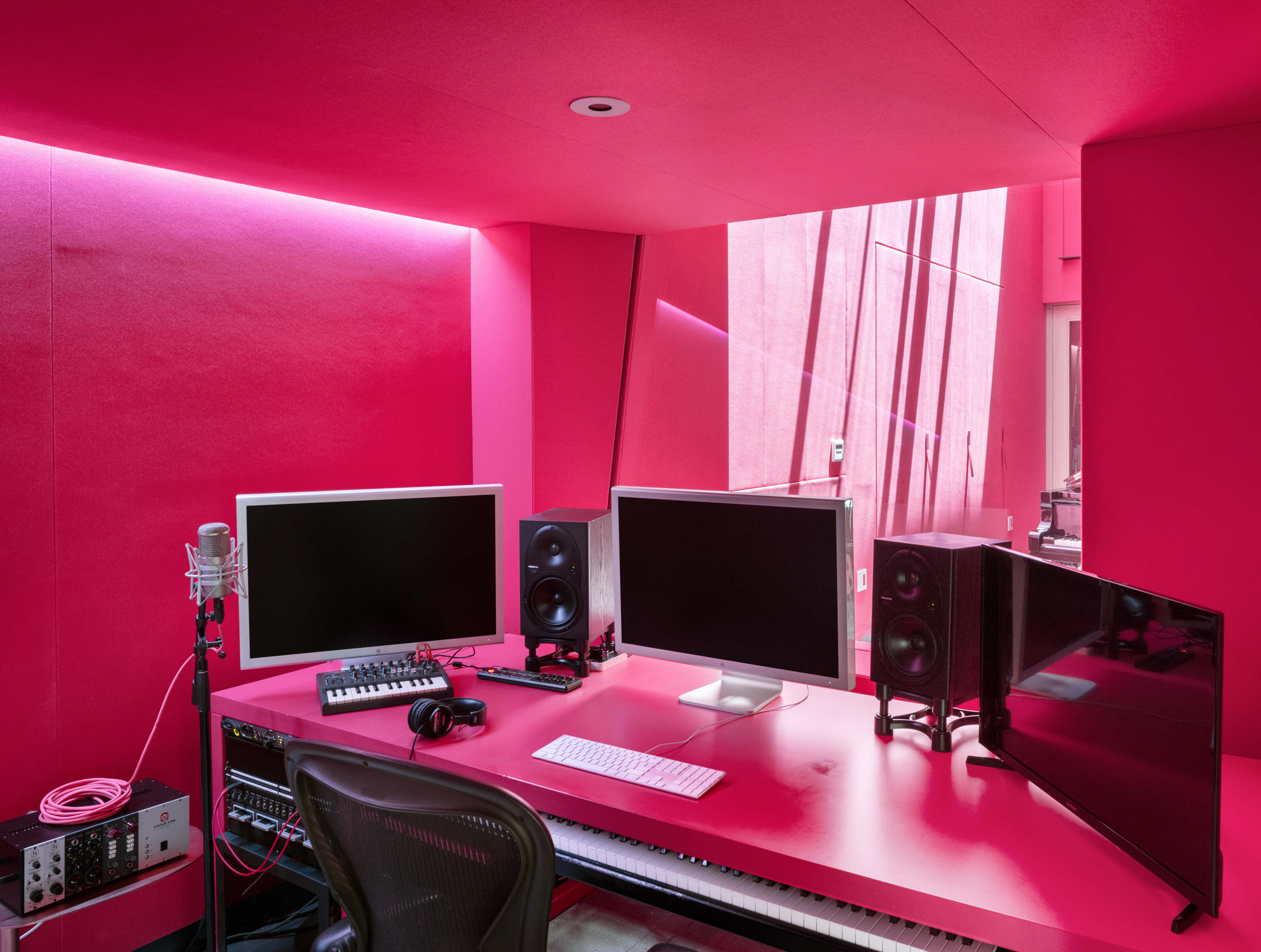 The image shows a music studio featuring pink walls, two computer monitors, speakers, a microphone, and a keyboard set up on a pink desk.