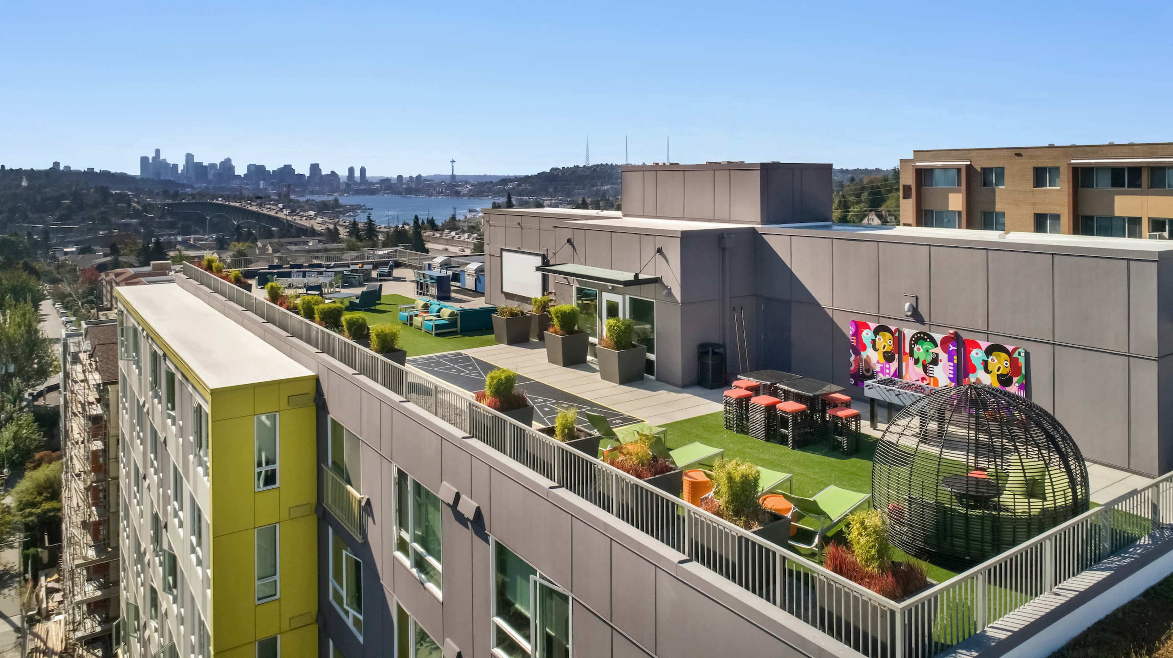 A rooftop garden with seating areas, colorful artwork on the wall, and a view of a city skyline in the background.