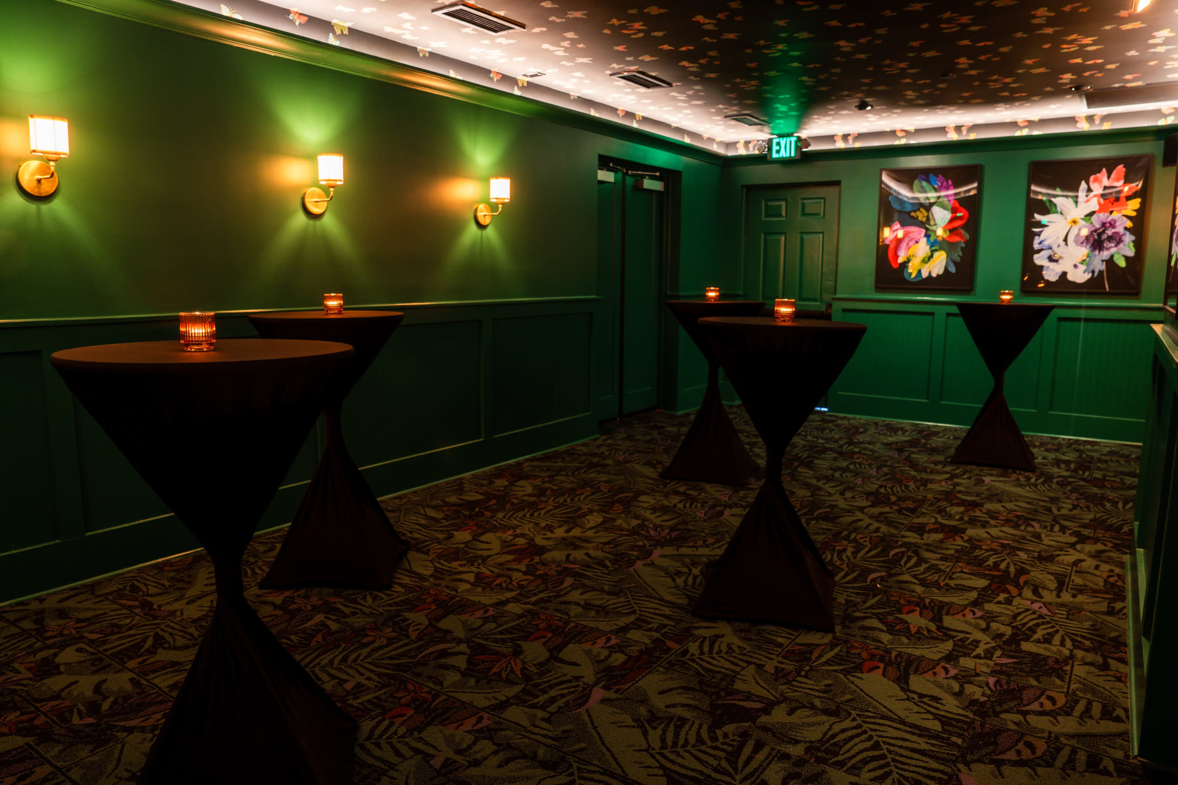 The image shows a dimly lit hallway with green walls, featuring three tall tables with black tablecloths and candles, and flower paintings on the wall.