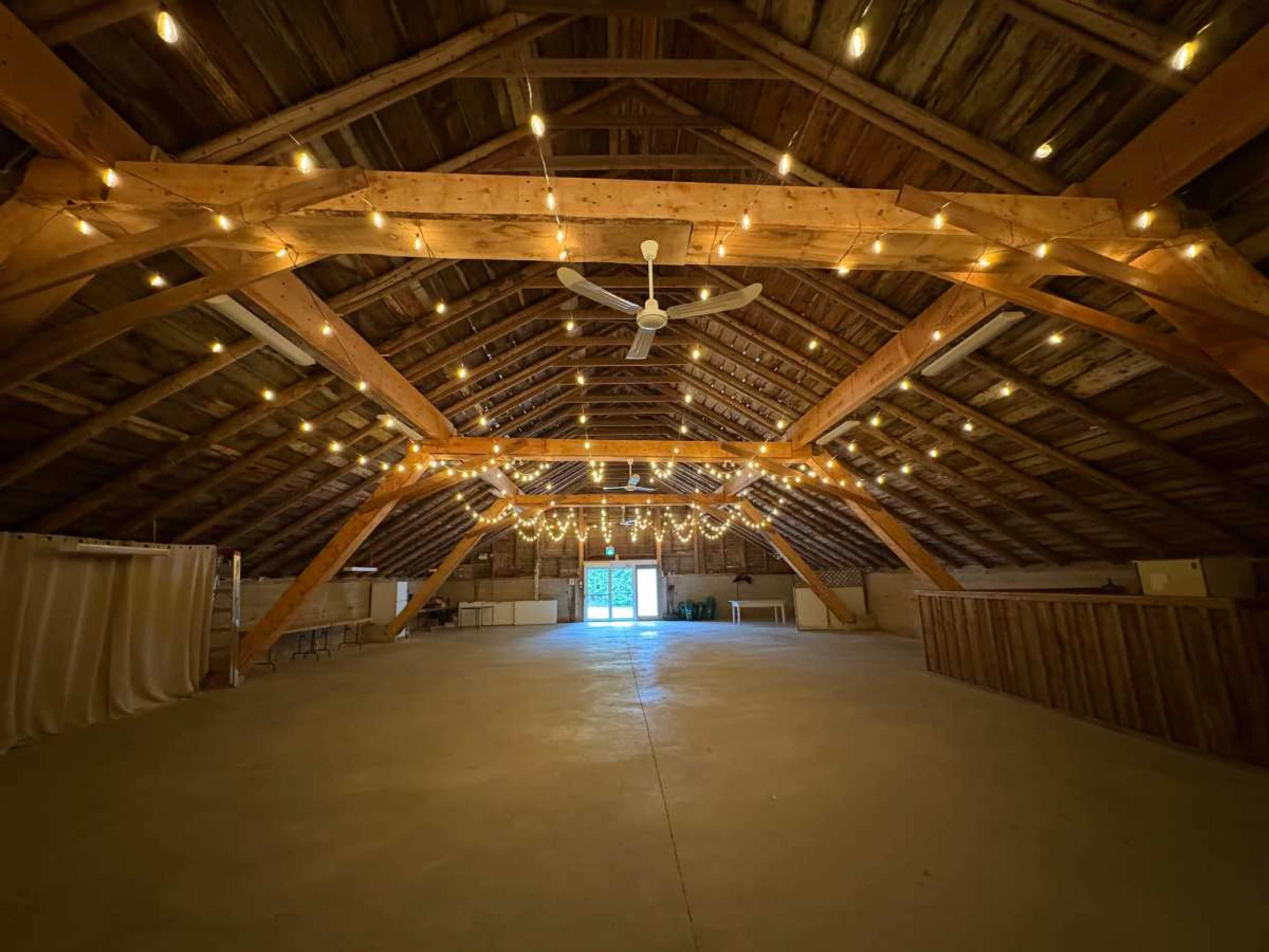 The image shows a spacious, rustic barn interior with wooden beams, string lights overhead, and an open entrance at the far end.