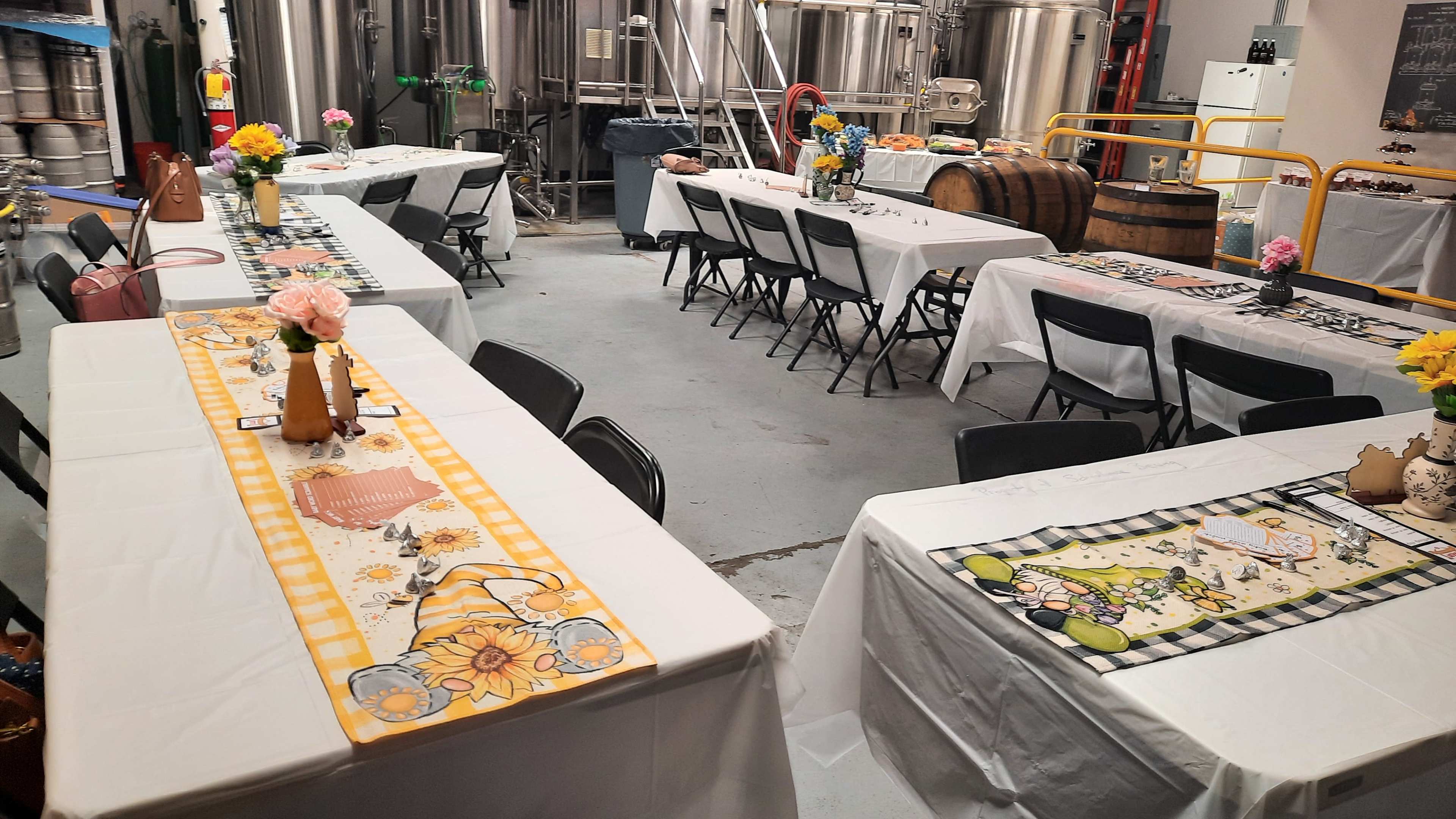 The image shows a room set up for an event, featuring several tables covered with white tablecloths, decorated with floral centerpieces, and surrounded by black chairs, with brewing equipment visible in the background.