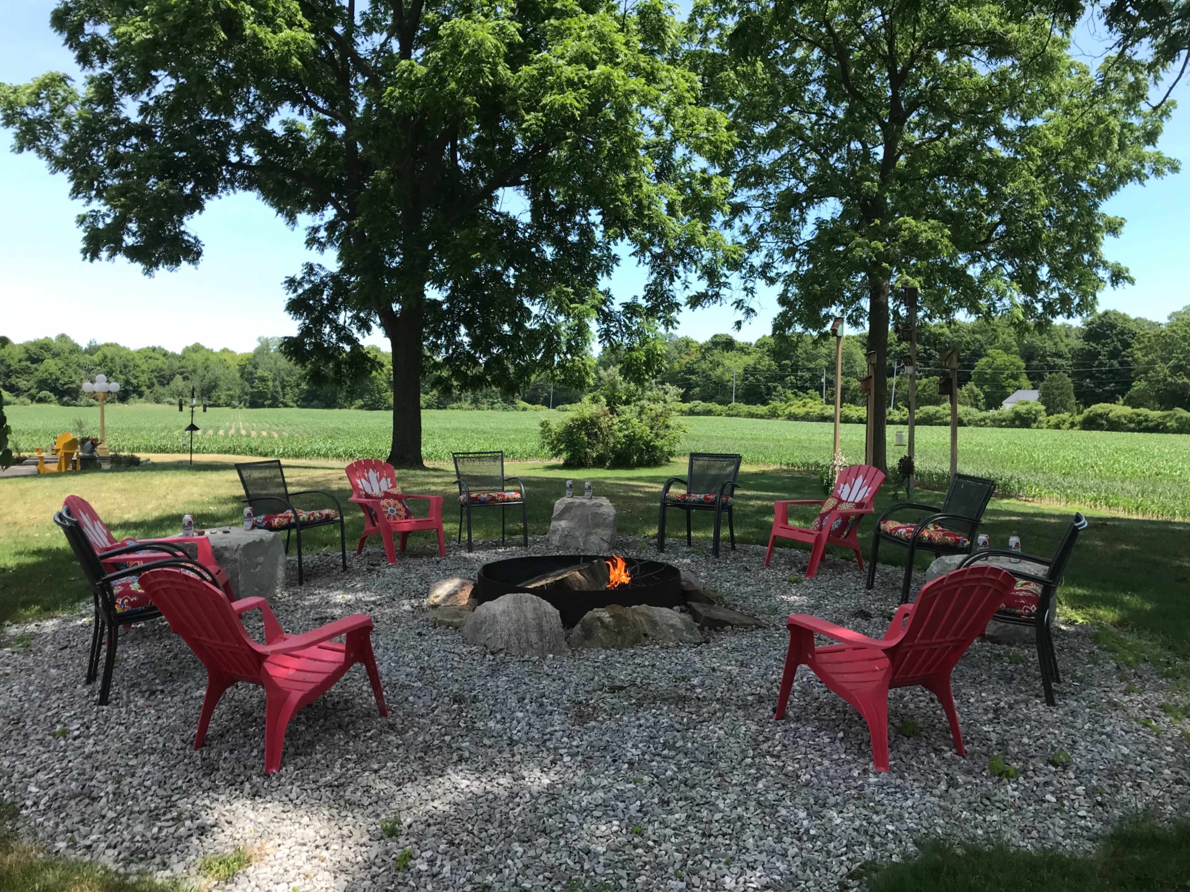 A stone fire pit surrounded by pink chairs on gravel, set in a grassy area with trees and a field in the background.