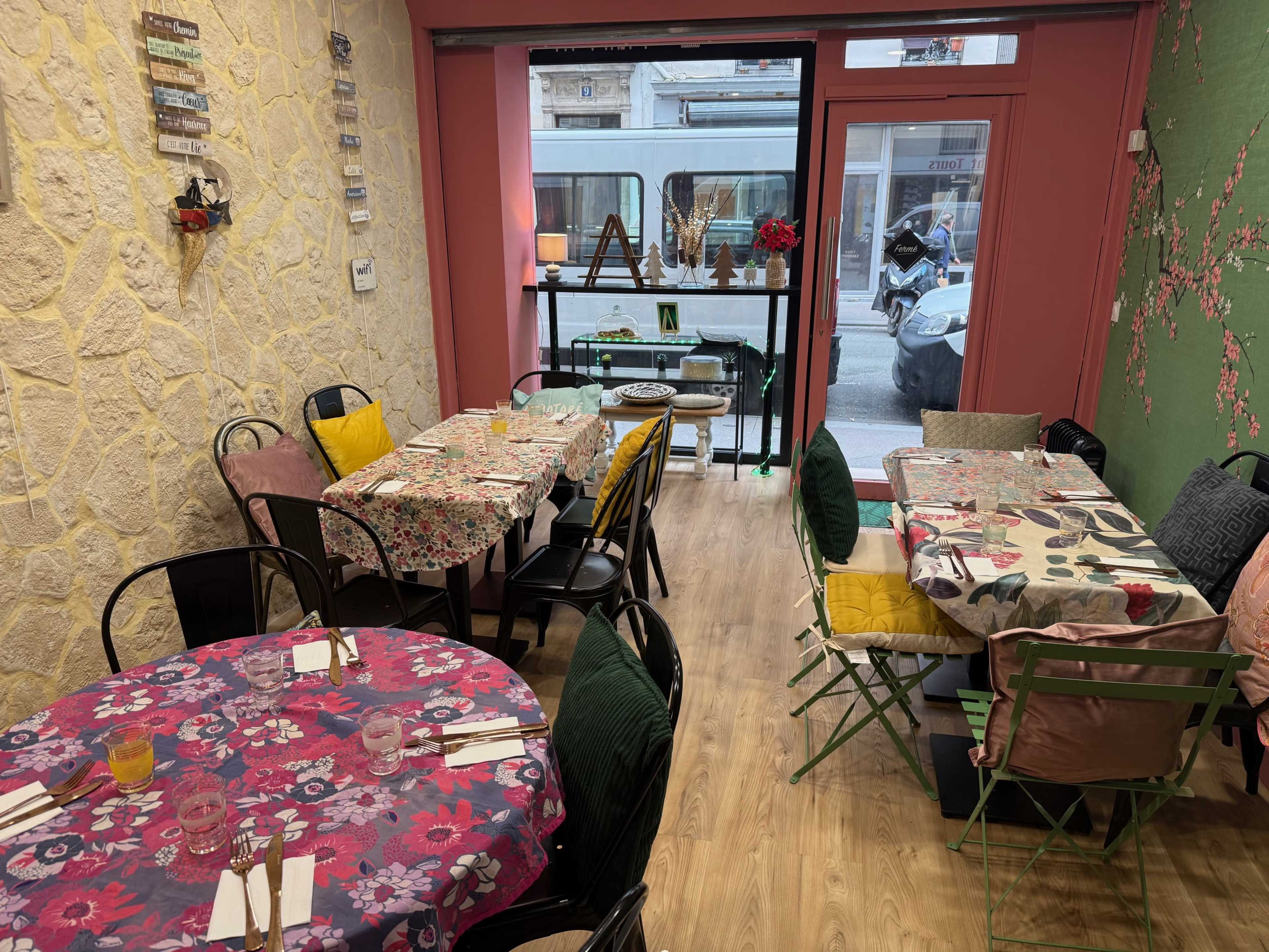 The image shows a cozy café interior with several tables, each covered in colorful tablecloths, and a window overlooking the street.