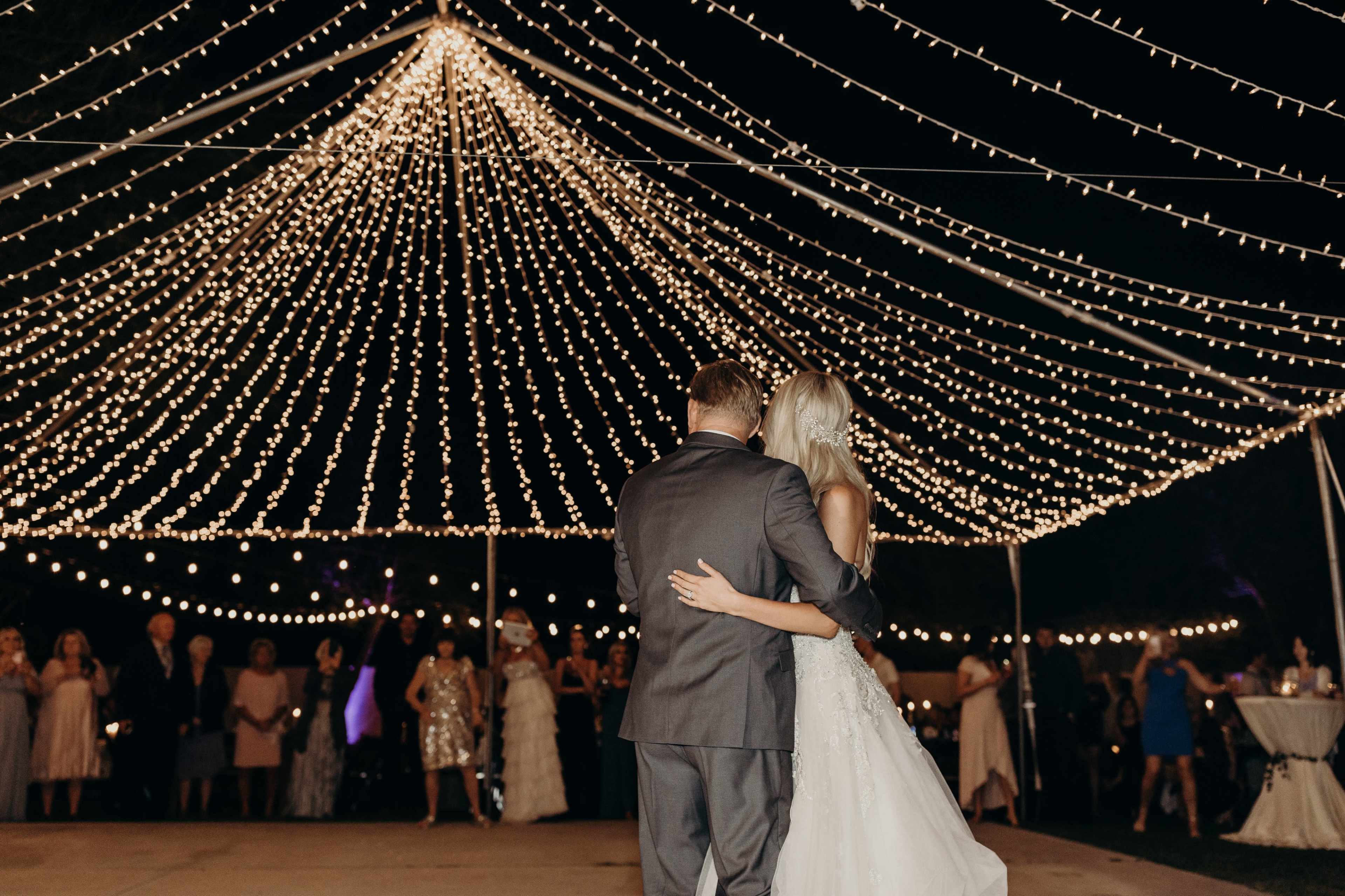 A couple dances under a canopy of lights at an outdoor wedding reception.