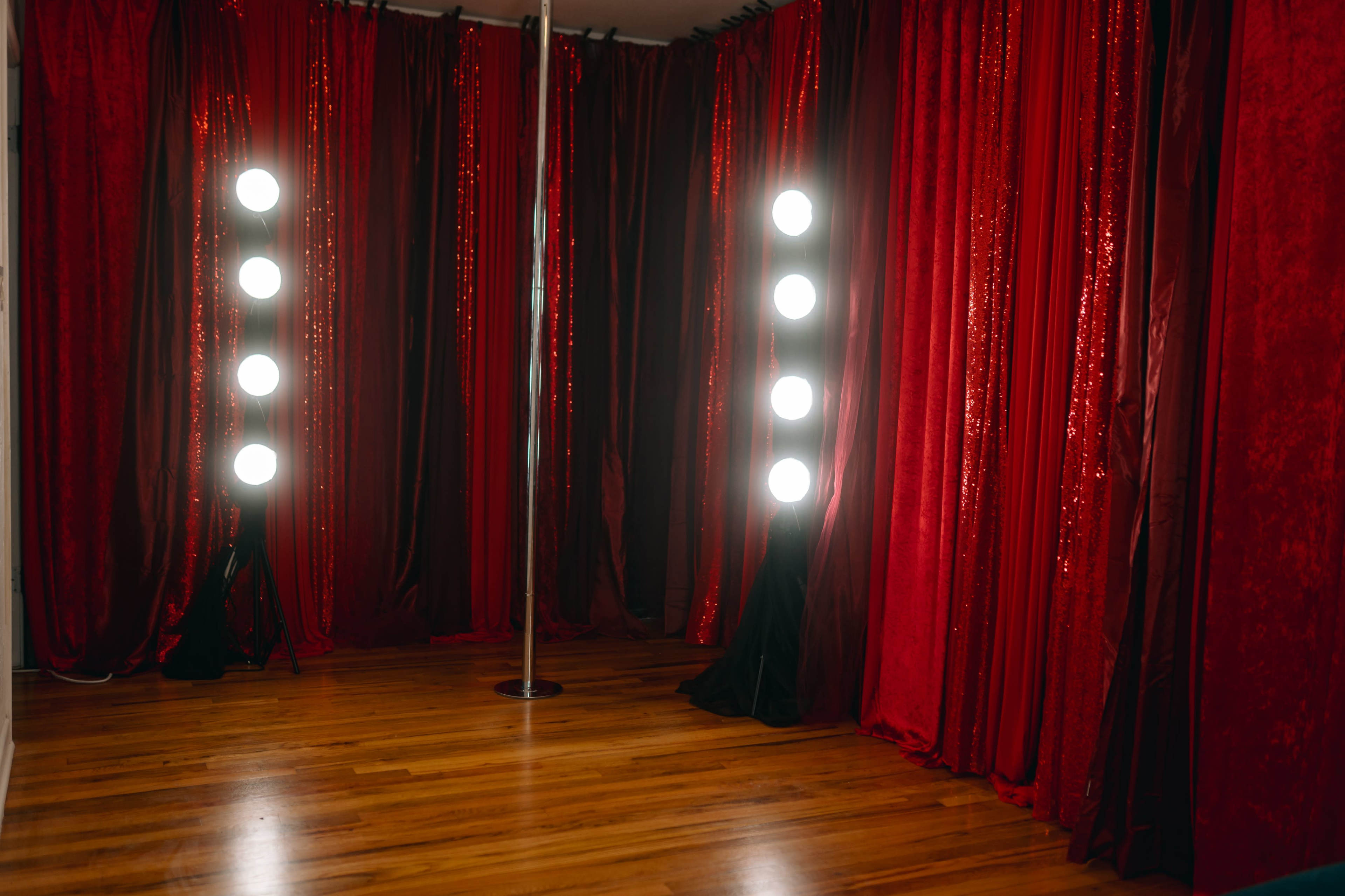 The image depicts a dance studio featuring a polished wooden floor, a metal pole in the center, and red velvet curtains illuminated by bright round light fixtures.