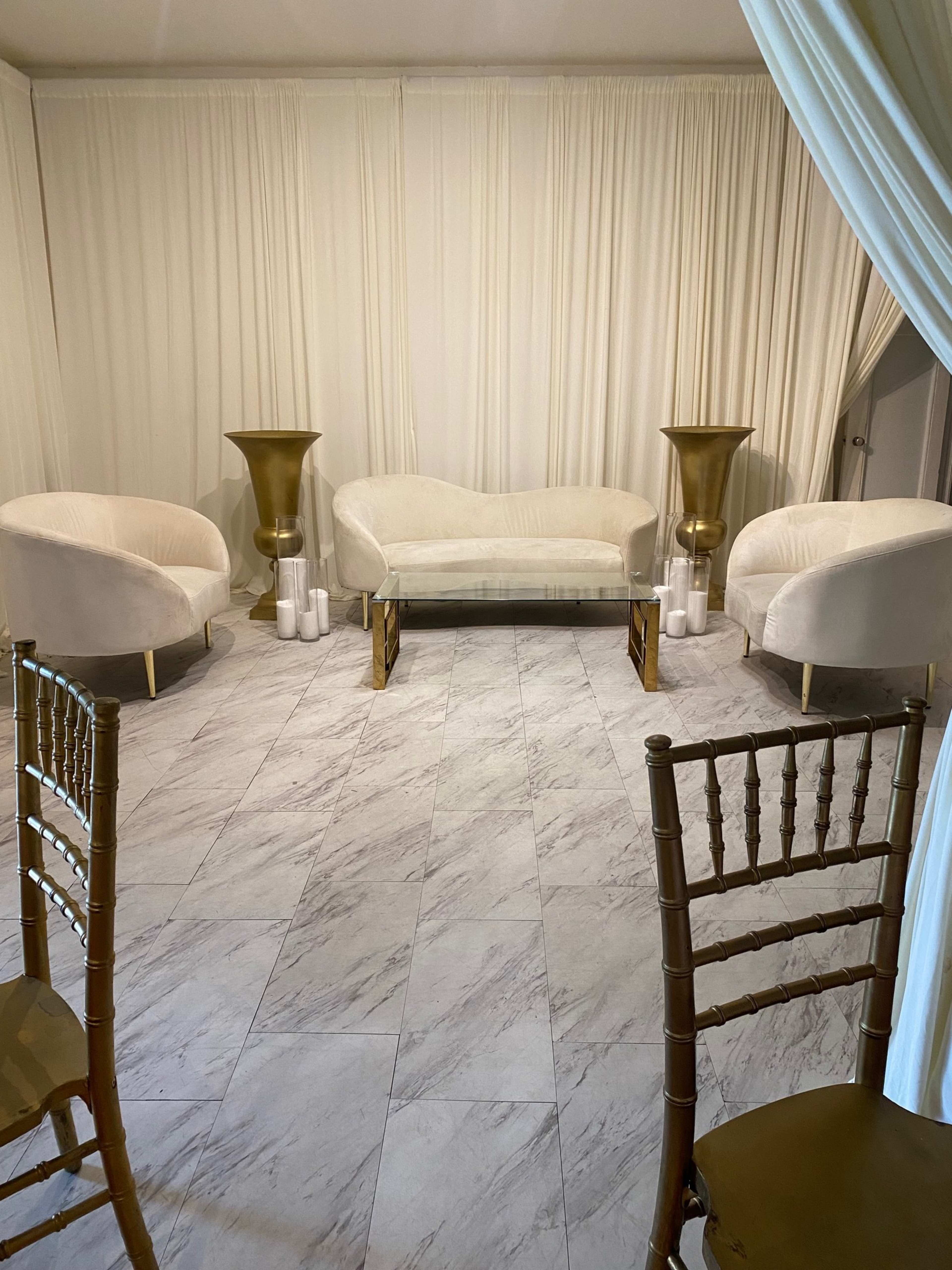 The image shows a small, elegantly arranged lounge area with a light-colored sofa and two matching chairs, flanked by gold-tone planters and candles on marble flooring.