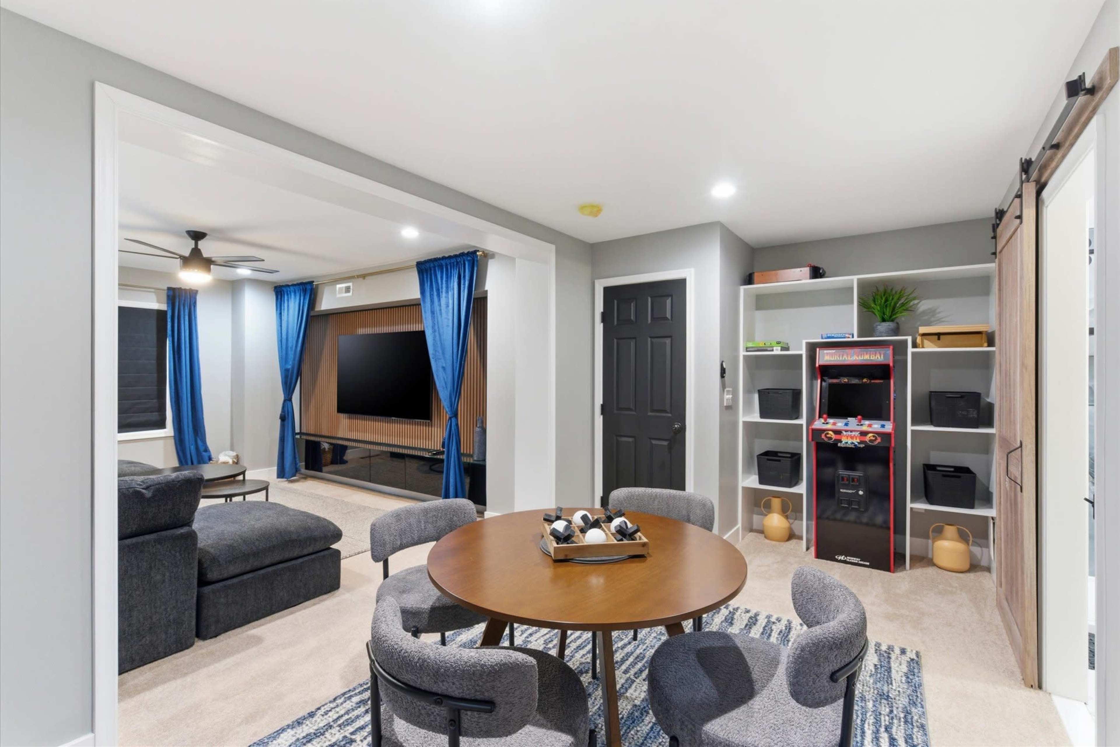 A cozy indoor space features a round table surrounded by four chairs, with a living area in the background that includes a television and shelves.