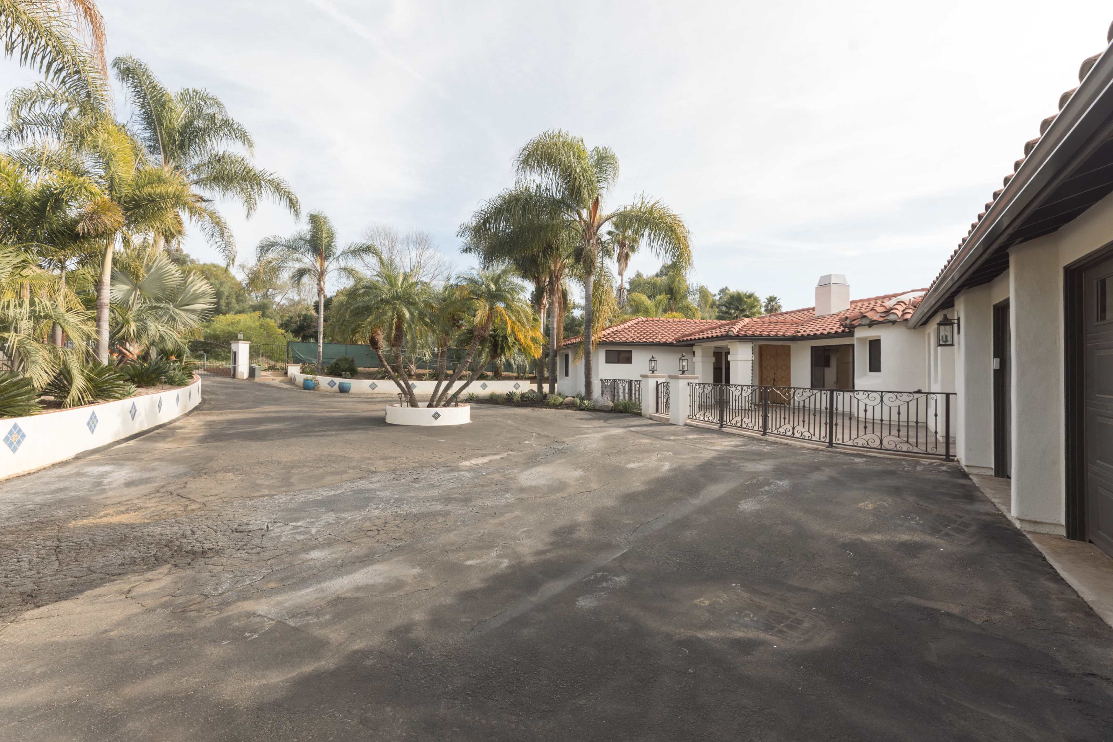 The image shows a driveway leading to a white stucco house with a red-tiled roof, surrounded by palm trees and landscaped greenery.